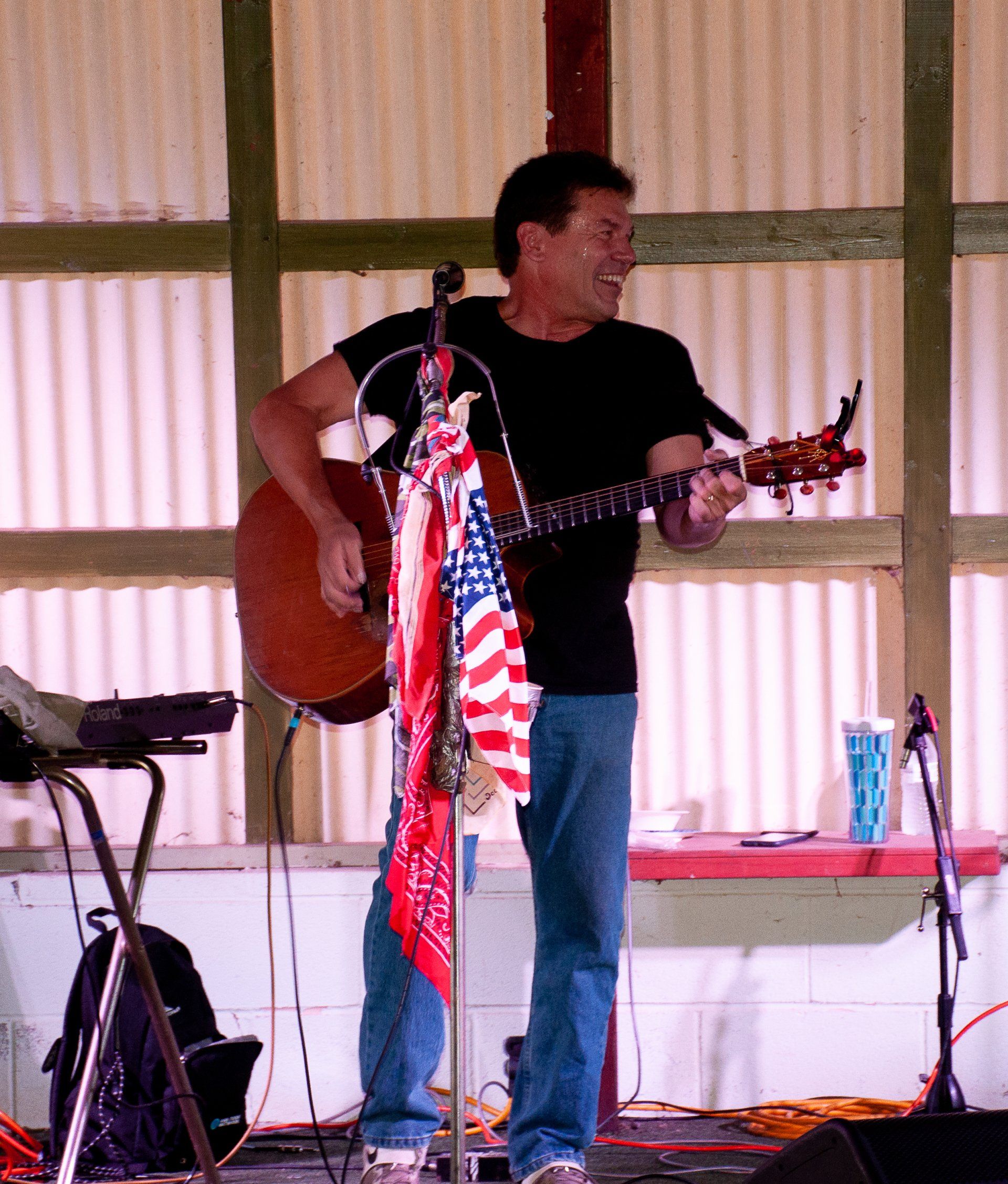 Man playing acoustic guitar on a small indoor stage with a microphone stand and striped backdrop