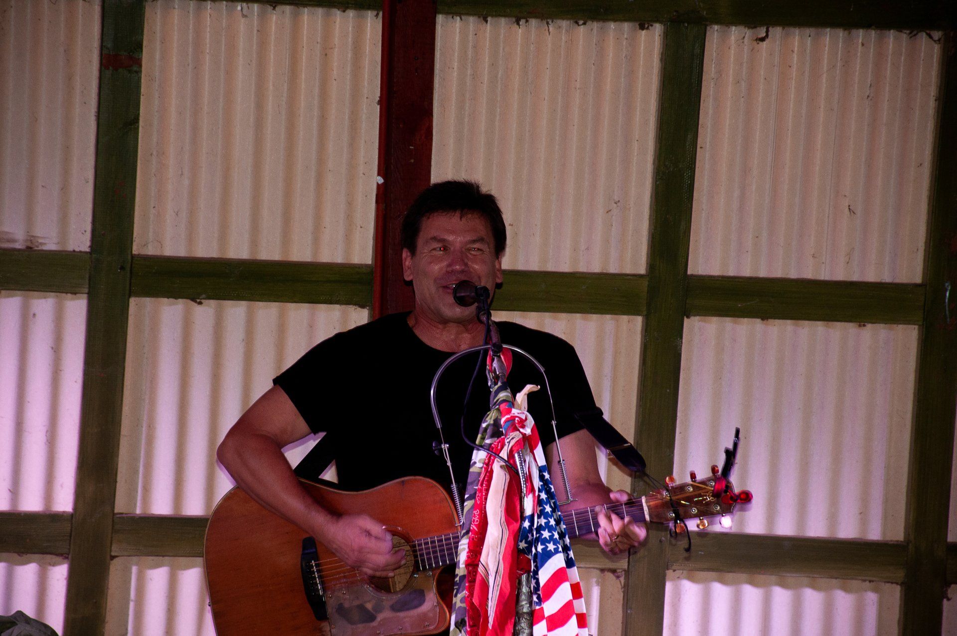 Man playing guitar and singing into a microphone on stage, with an American flag in front.