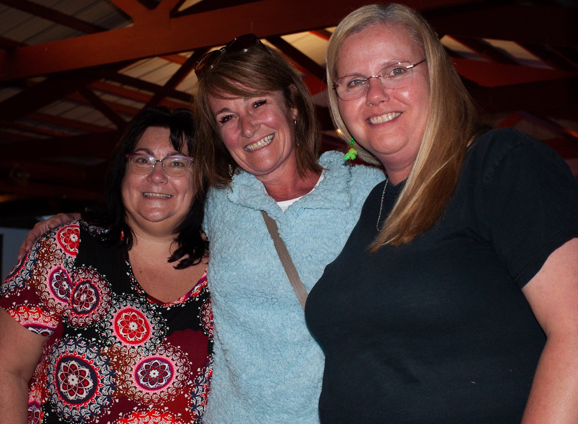 Three people smiling indoors, leaning together in a dimly lit room.