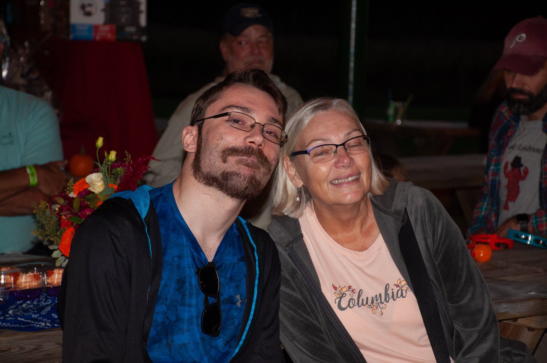 Two smiling people leaning together at a dimly lit indoor event, with flowers and people in the background