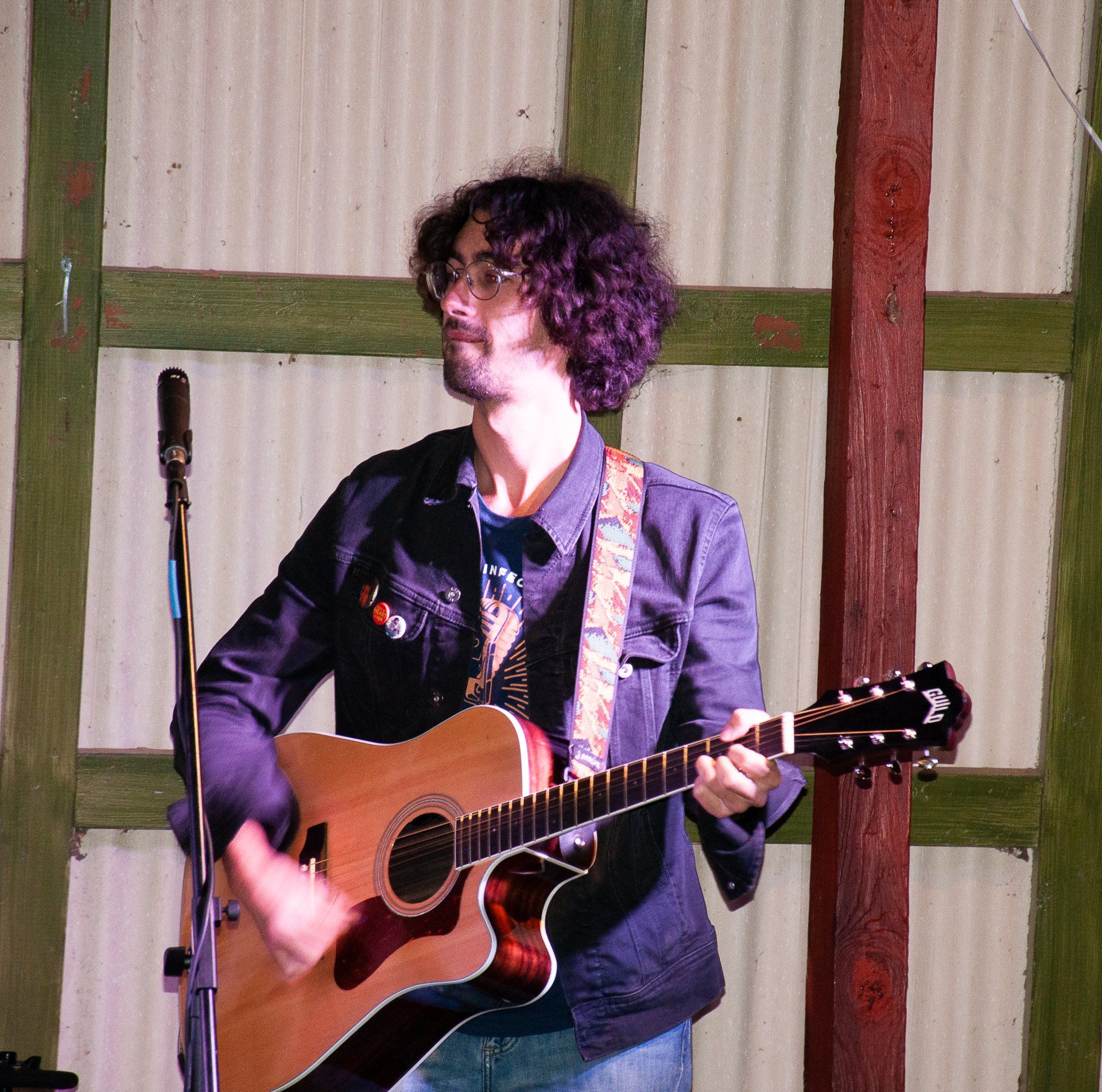 Musician singing and playing acoustic guitar onstage beside a microphone, wearing a dark jacket.