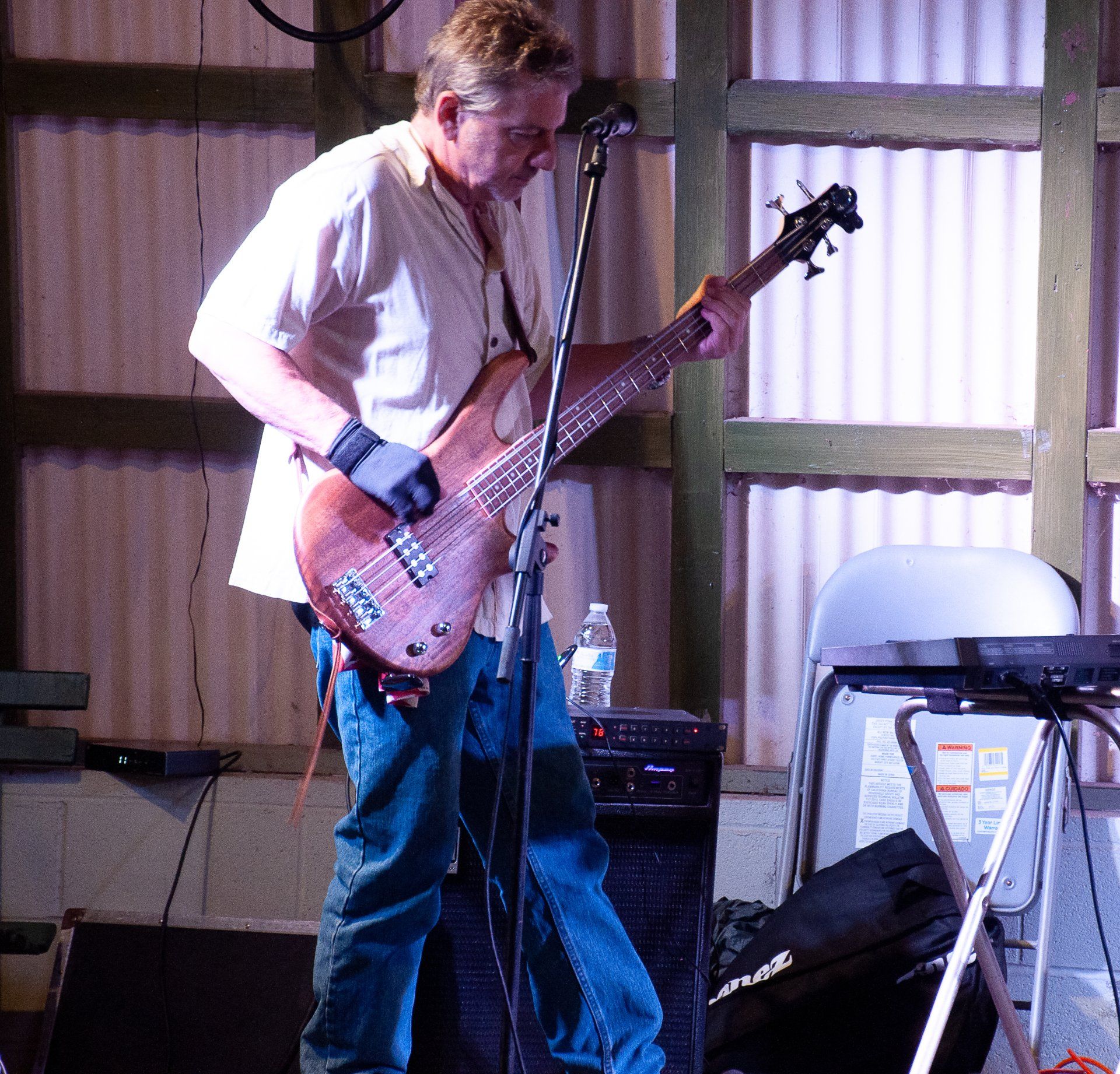 Musician playing bass guitar on a small indoor stage, standing beside a microphone and speaker.