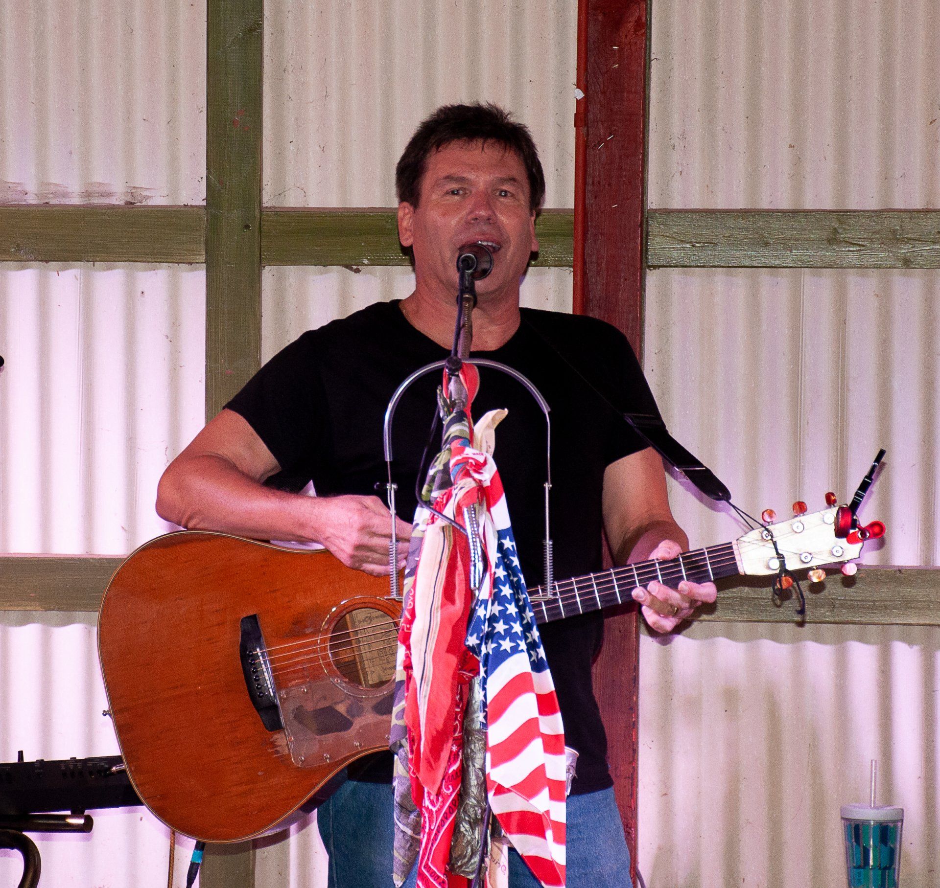 Singer playing acoustic guitar at a microphone, with an American flag draped on the stand.