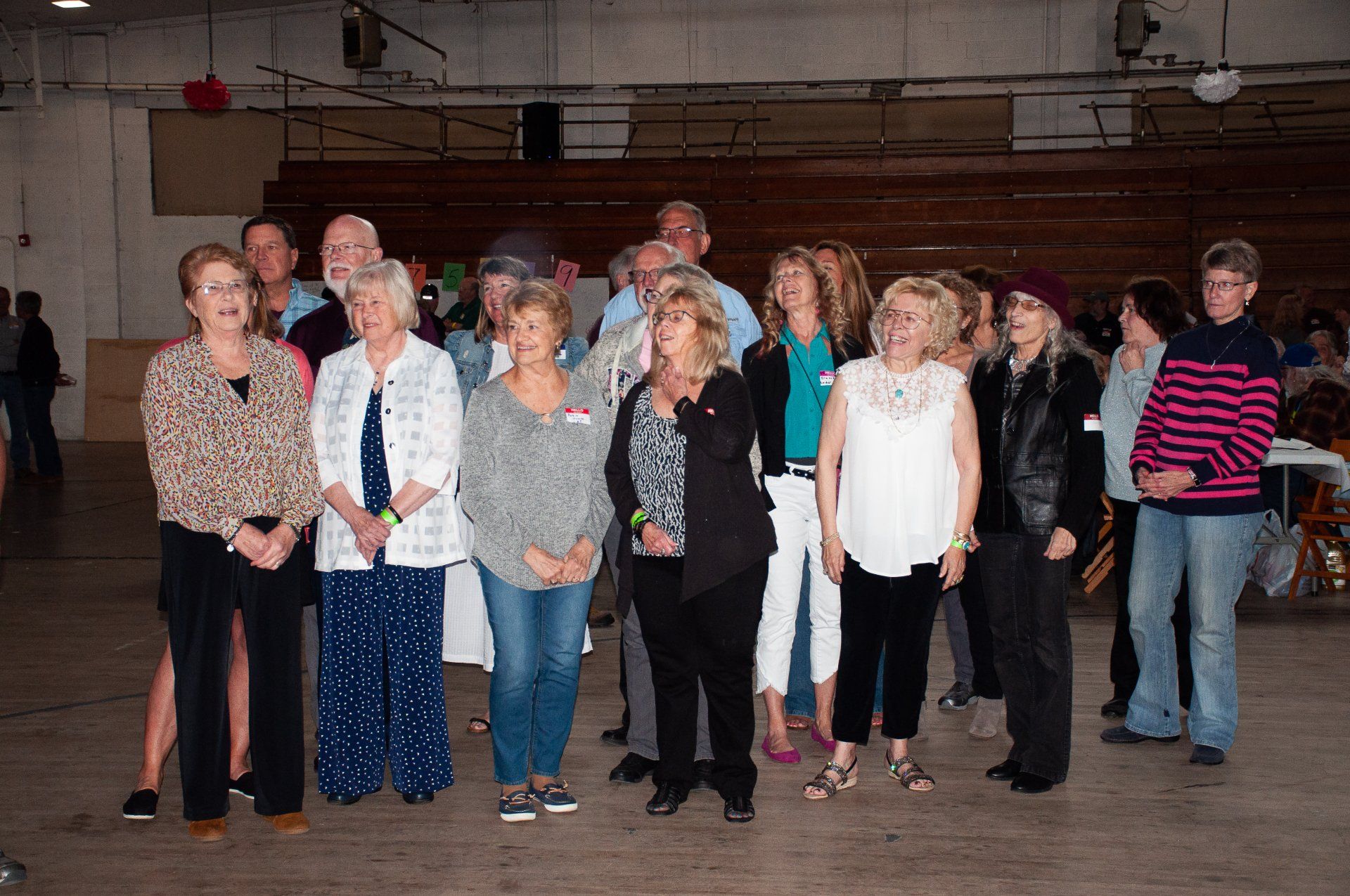 Group of people standing indoors in a large hall, facing the camera, some holding papers and wearing casual clothes.