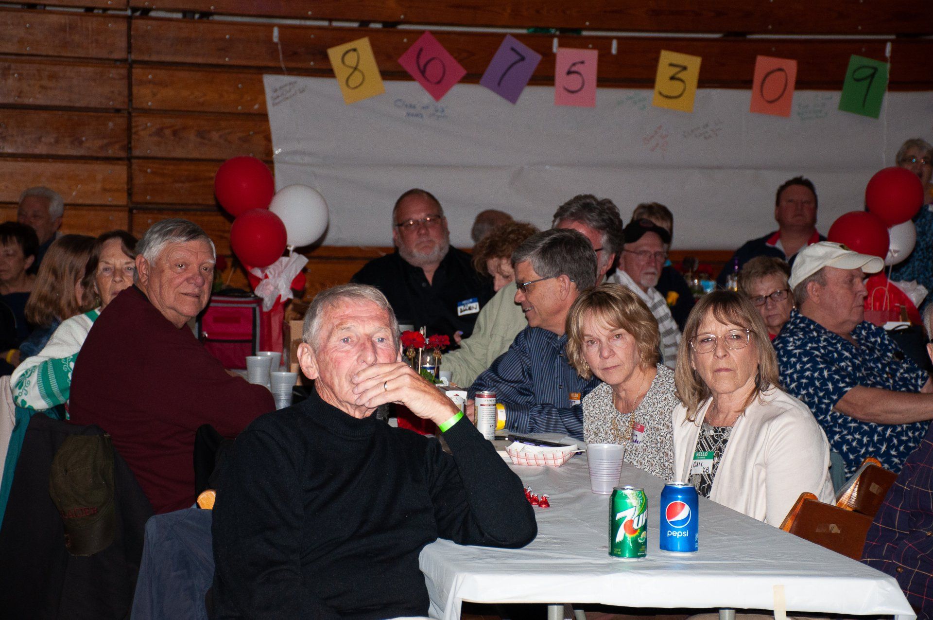 People seated at tables in a festive hall, with balloons, holiday decor, and drinks on the tables.