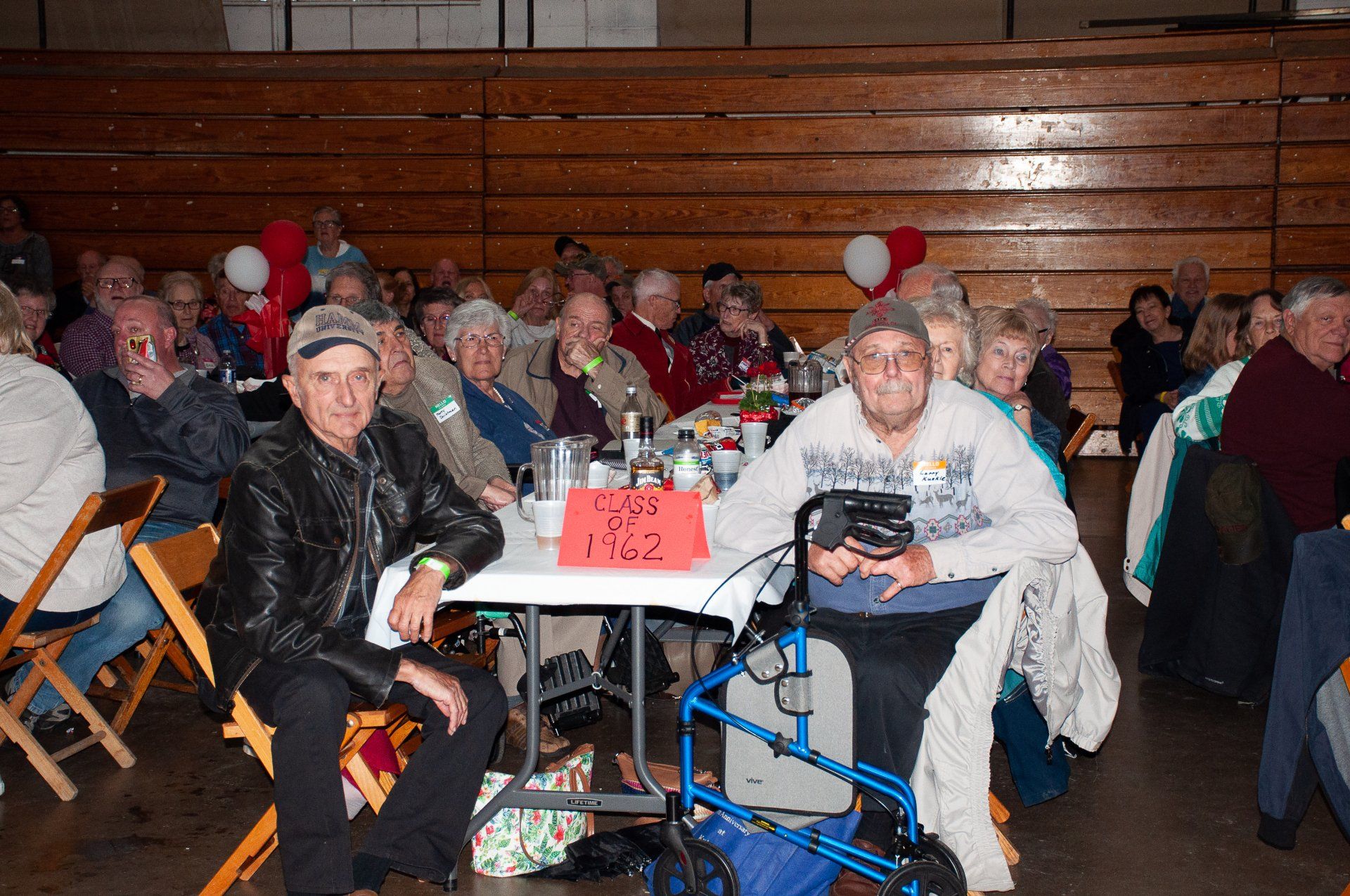 People seated at folding tables in a gym, with balloons and a sign on the front table.