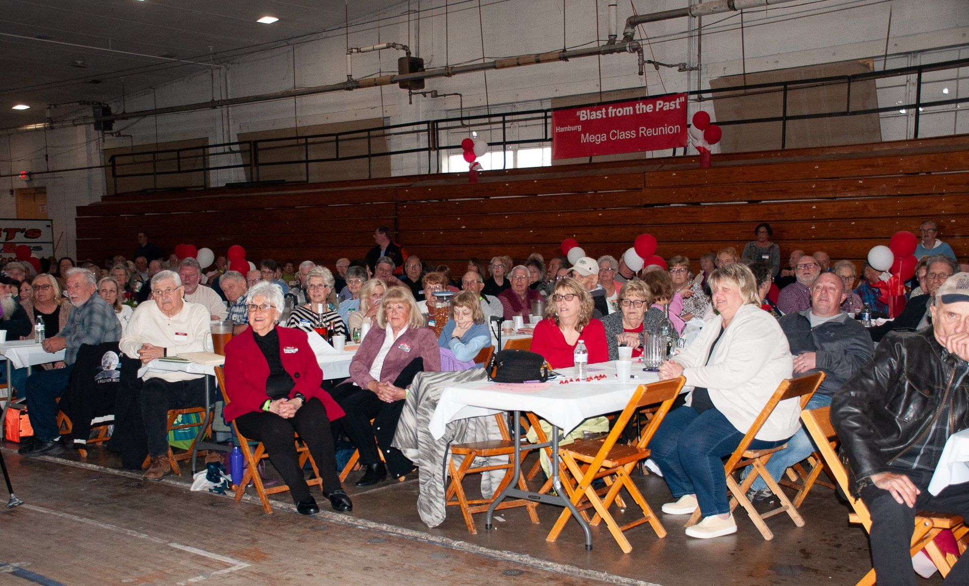 Crowd seated at folding tables in a gymnasium, listening at a community event with red banners in the background