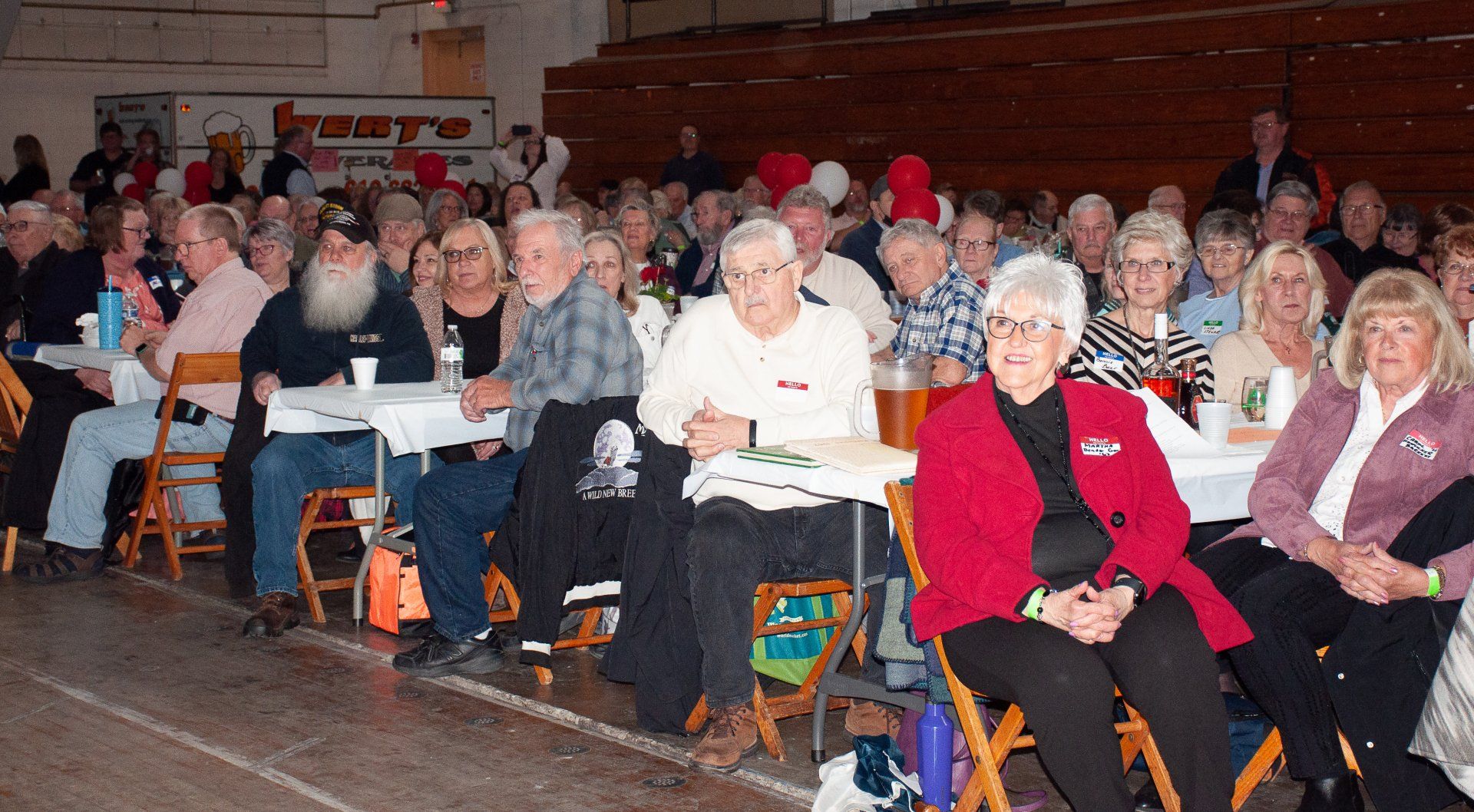 Audience seated in a gymnasium, many holding papers and wearing winter clothing.