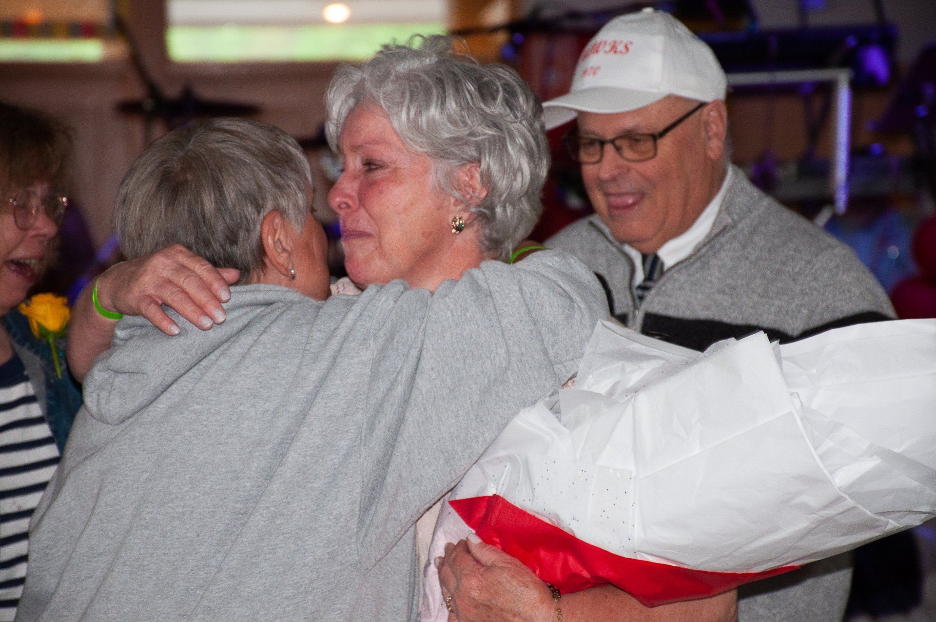 Two people embrace while a man smiles nearby holding a red-and-white gift bag.