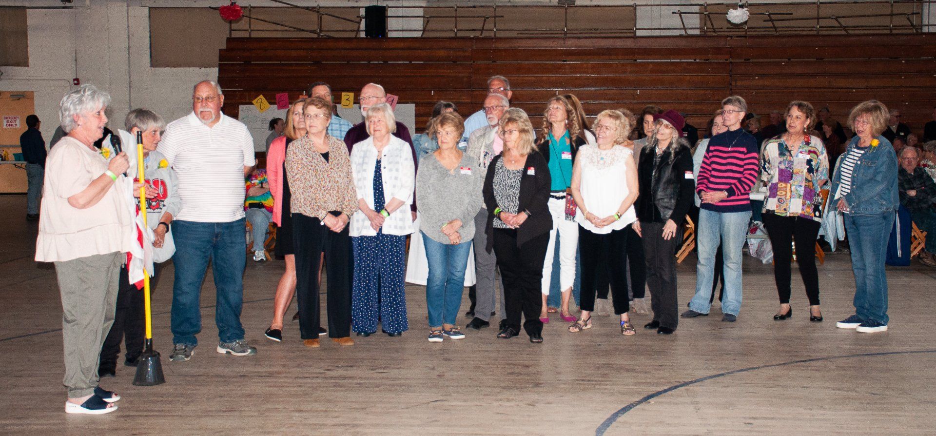 Group of people standing in a gymnasium, posing on a wooden floor in front of bleachers.