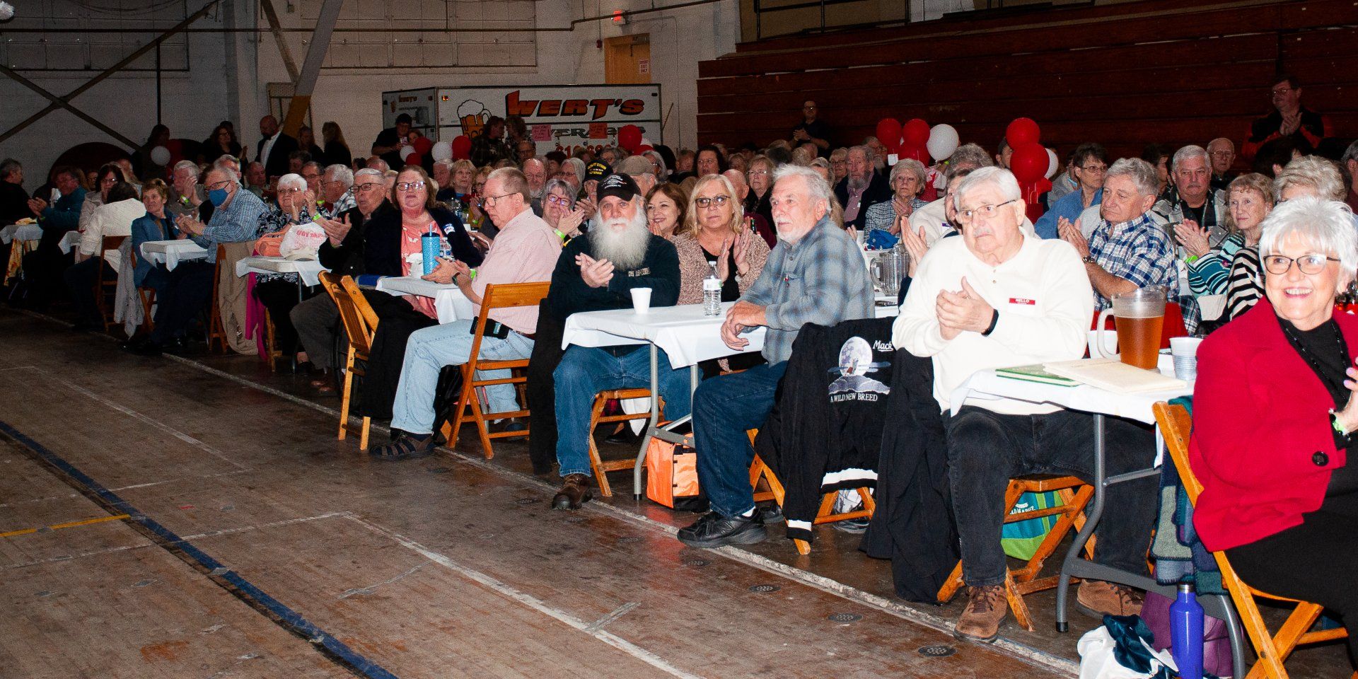 Audience seated on gym floor in a large hall, facing a stage or event off-camera