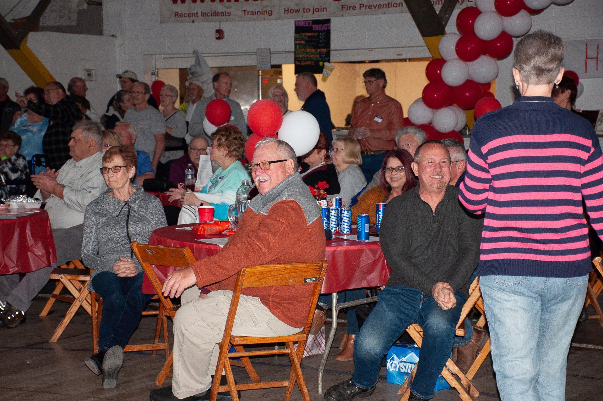 People seated at tables in a festive hall decorated with red-and-white balloons during a gathering.