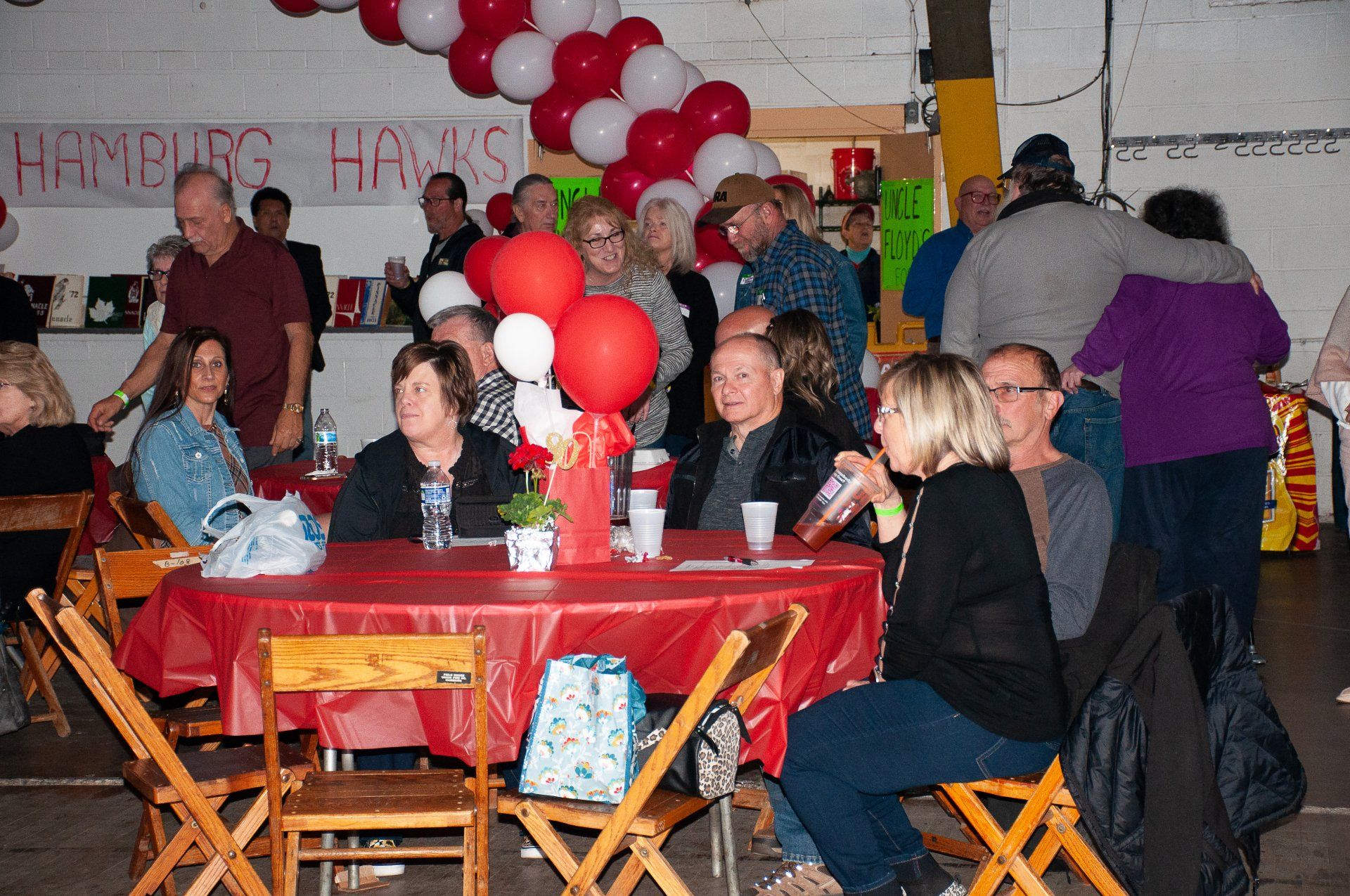 People seated at red-covered tables in a decorated indoor hall with red and white balloon arches