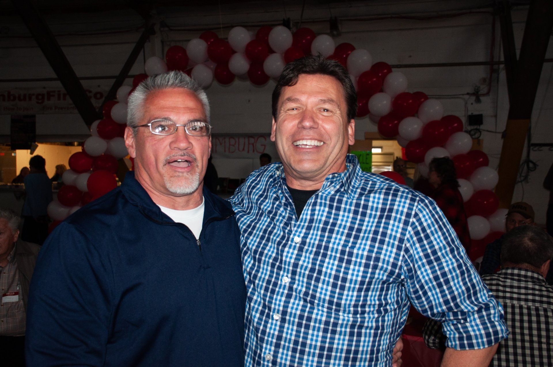 Two men smiling at an indoor party with red and white balloons in the background
