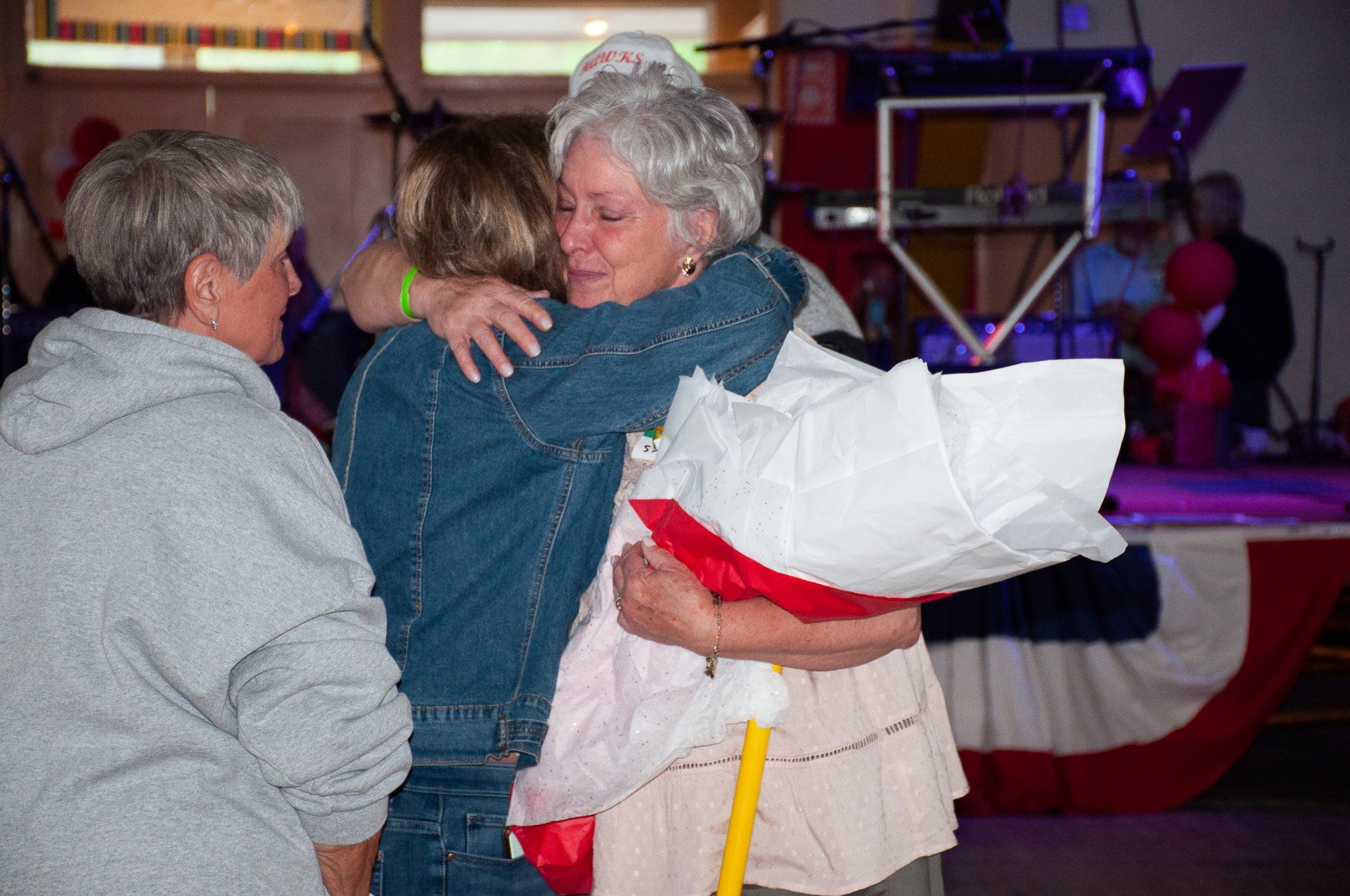 Two people hugging at a party, with a woman holding a white-and-red wrapped bouquet.