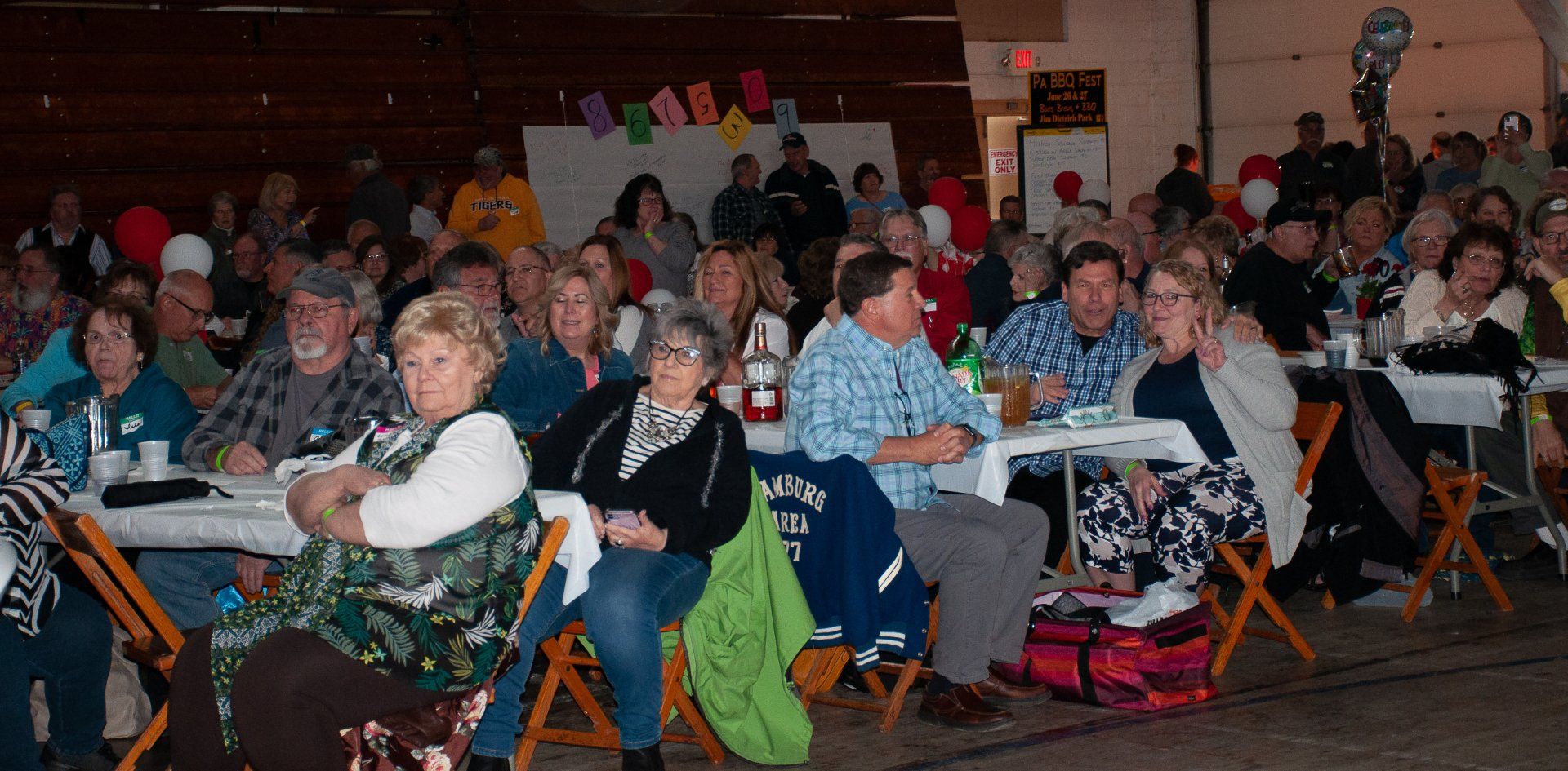 Crowded indoor gathering with seated audience members at tables, watching an event in a dimly lit hall