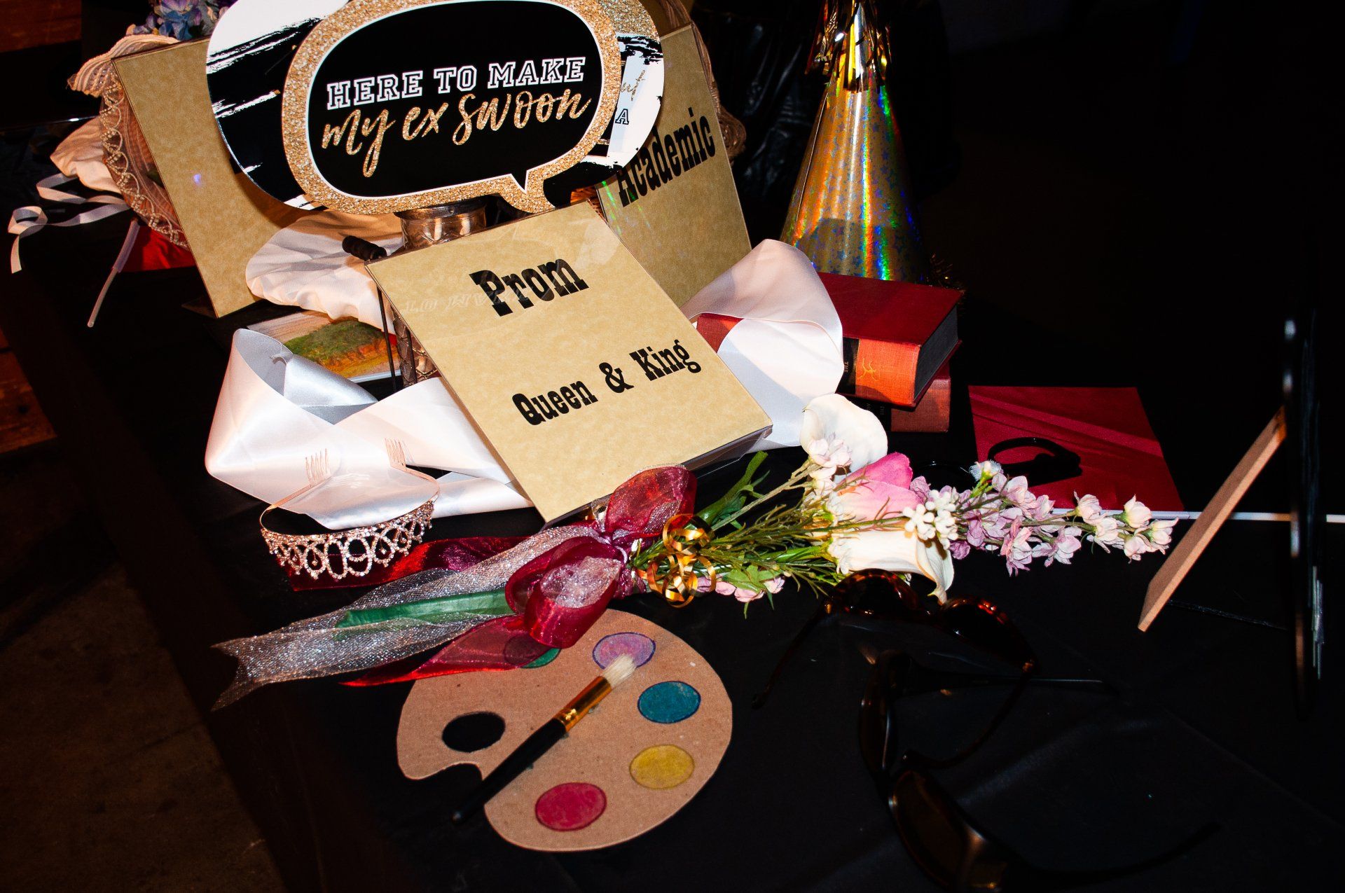 Craft supplies and party decor with brushes, paints, flowers, and a palette on a dark table.