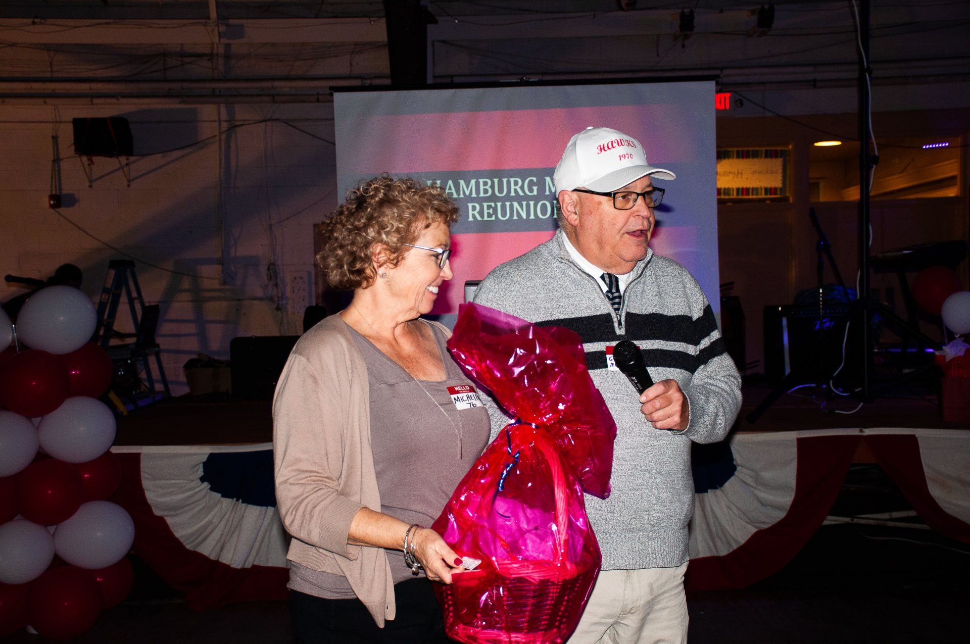 Two people holding a large red heart balloon at an indoor event with stage lights and a presentation screen.