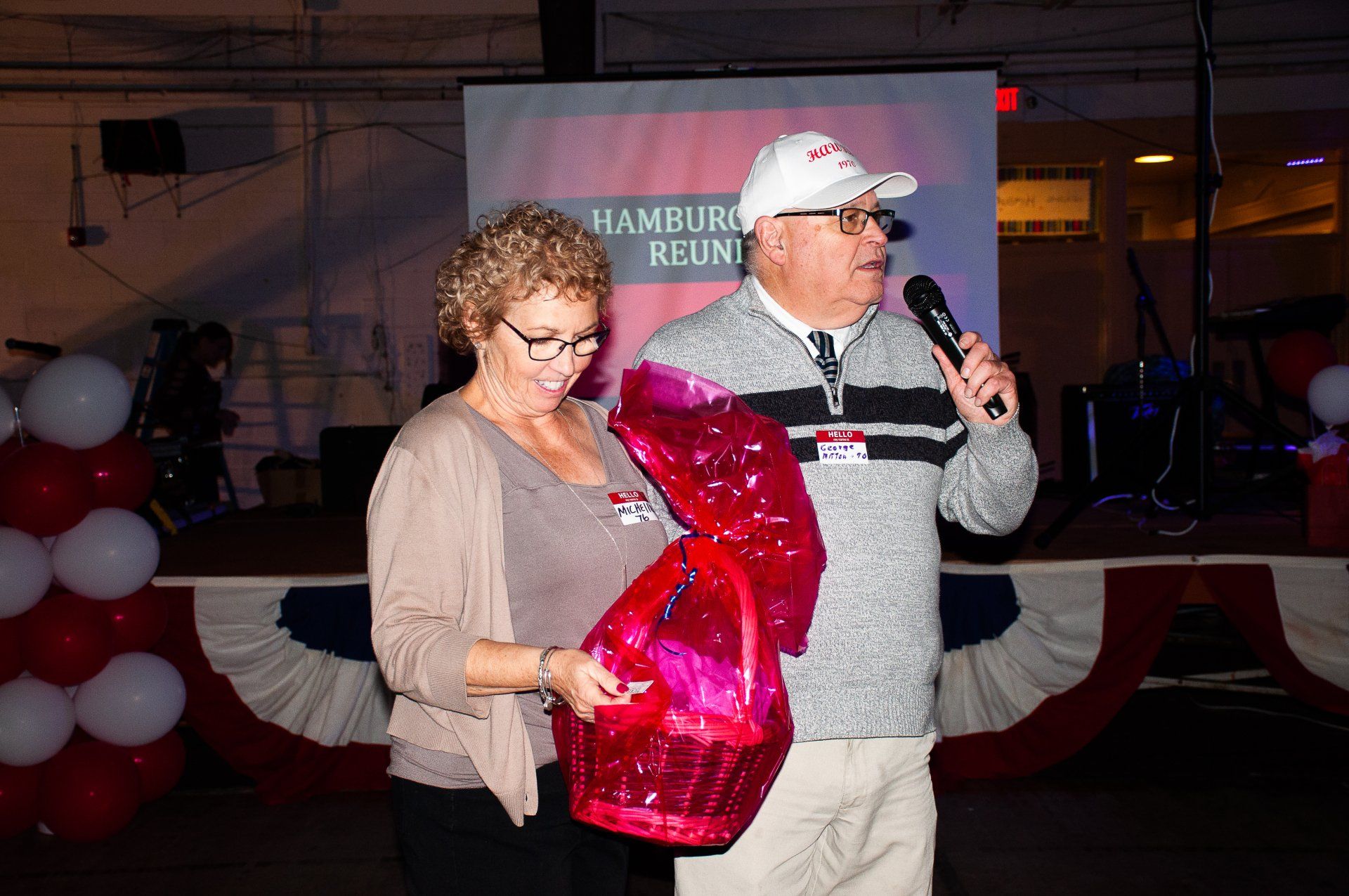 Two people on stage, one holding a red gift bag while the other speaks into a microphone.
