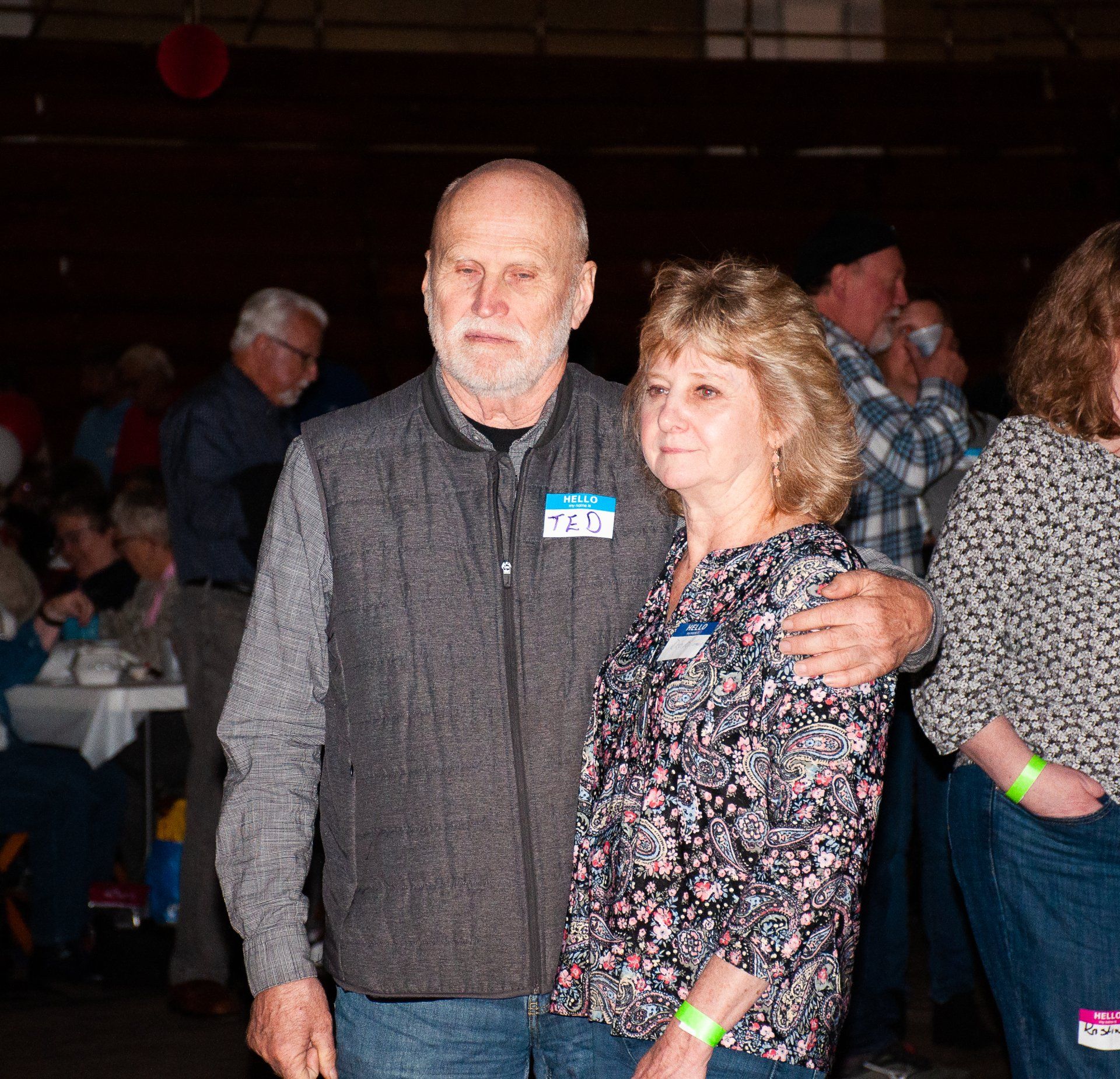 Two people posing together at an indoor event, wearing casual clothes and green wristbands.