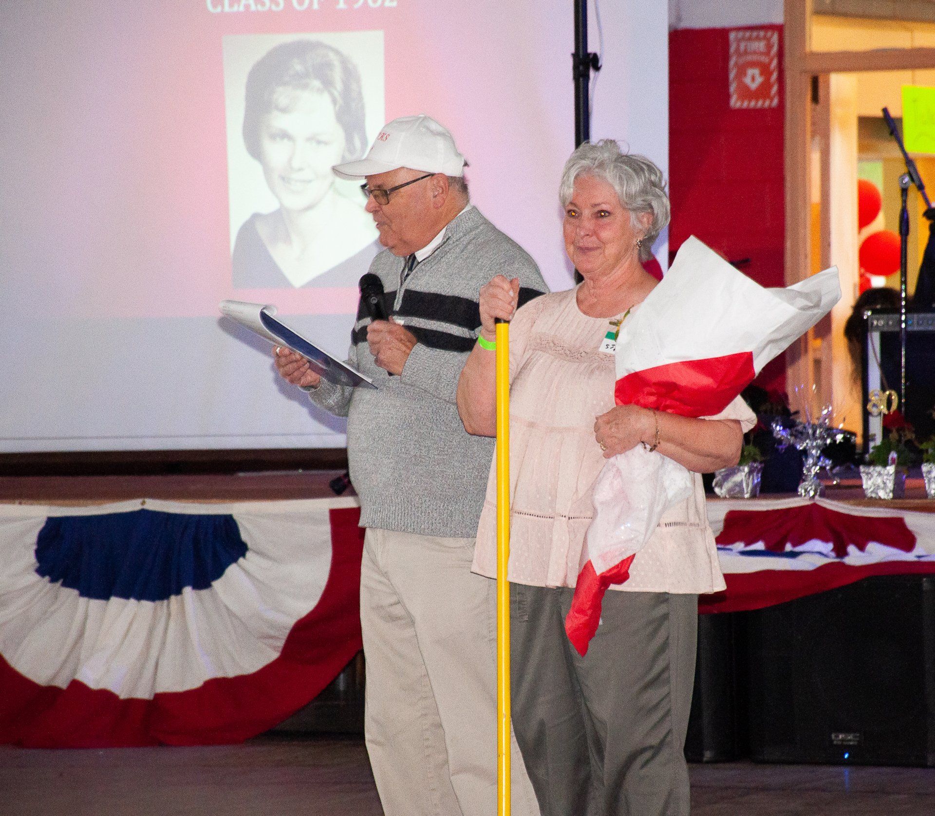 Two people on a decorated stage, one speaking from notes and the other holding a wrapped bouquet.