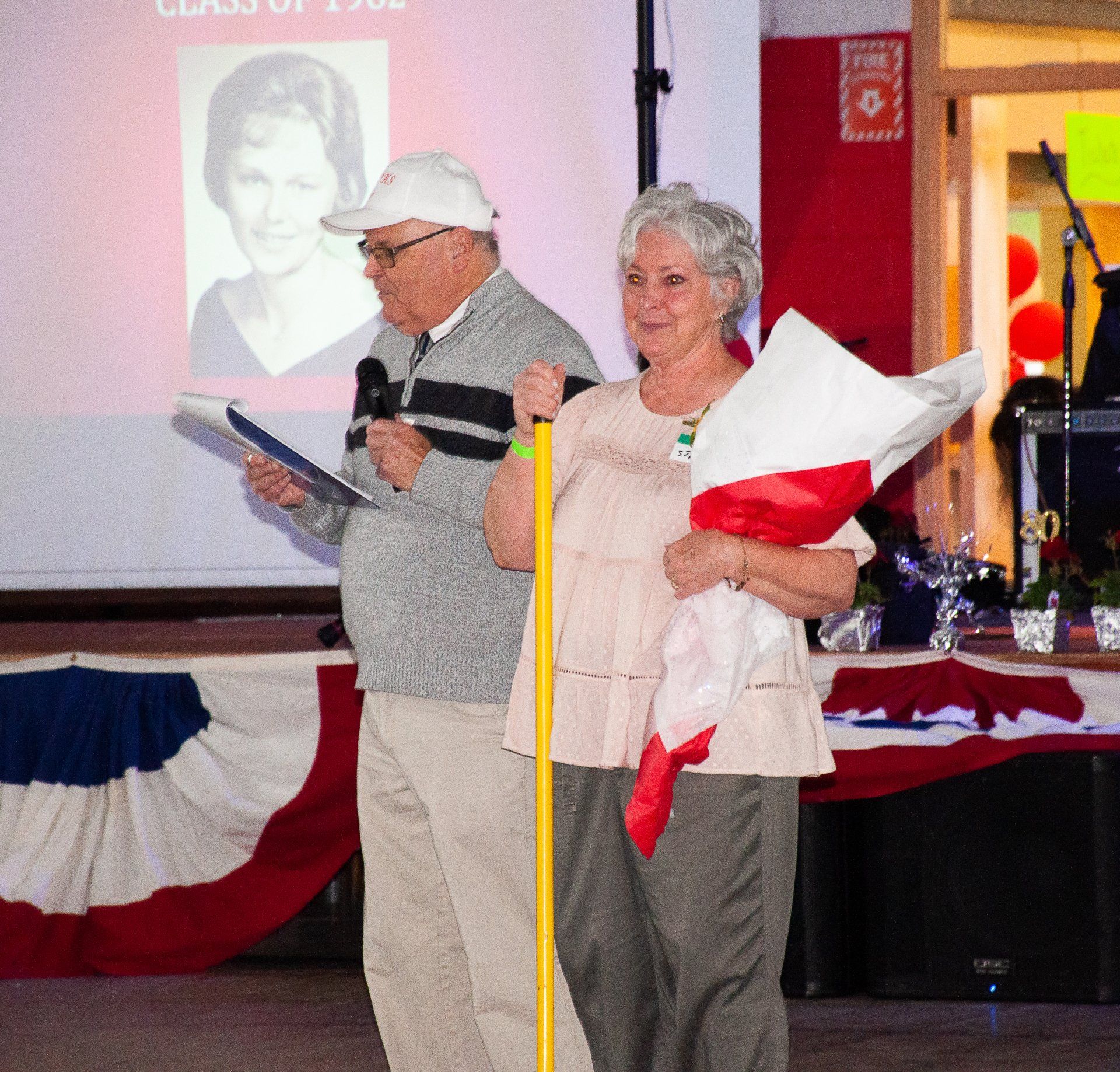 Two people at a podium in a decorated hall, one reading from notes and one holding a bouquet.