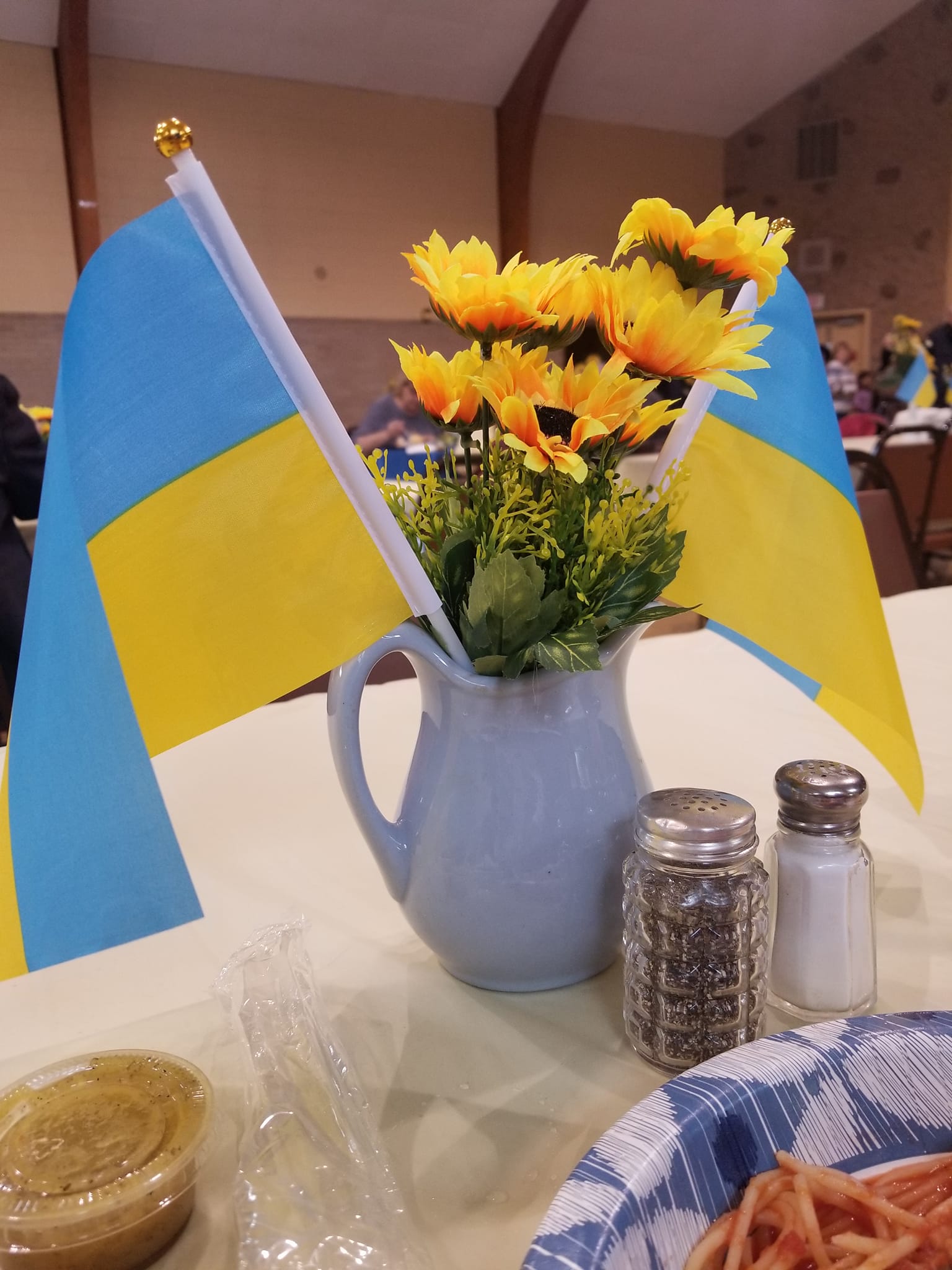 Small table with a sunflower vase, two blue-and-yellow flags, and condiments in a café-like setting