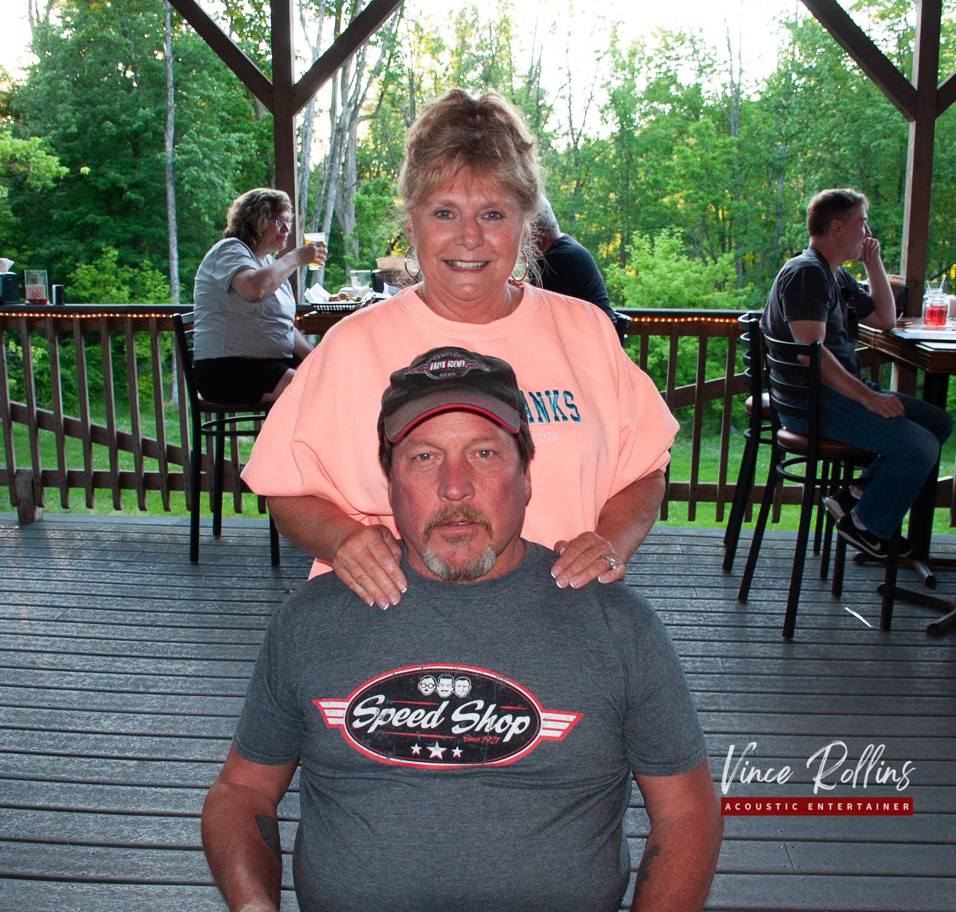 Smiling woman in peach shirt standing behind seated man in gray cap on a wooden deck.