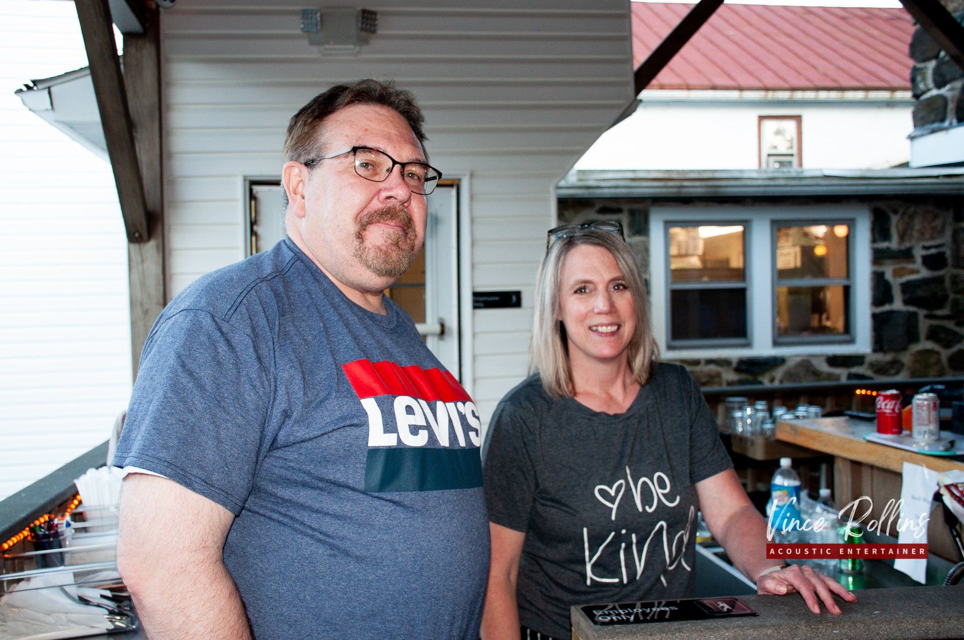 Two people standing on a porch beside a table with drinks and food, smiling at the camera.