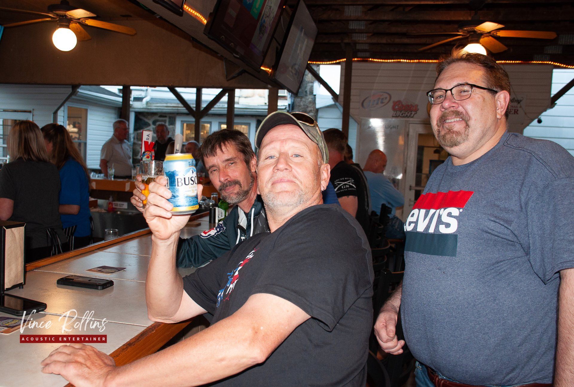 Three men in a rustic bar; one smiles while holding up a drink can.