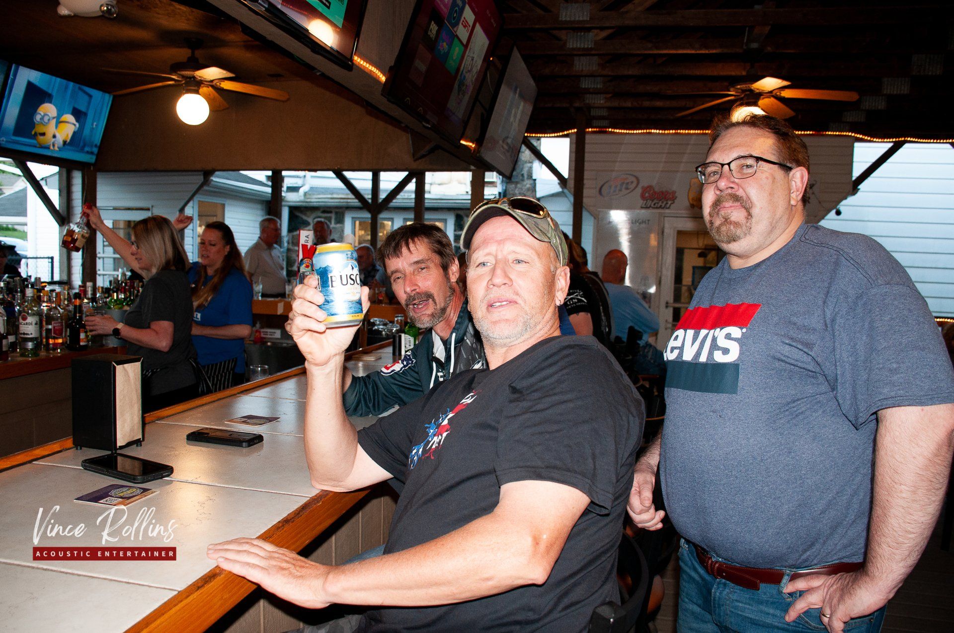 Three men at a bar counter, one holding a drink and smiling toward the camera.