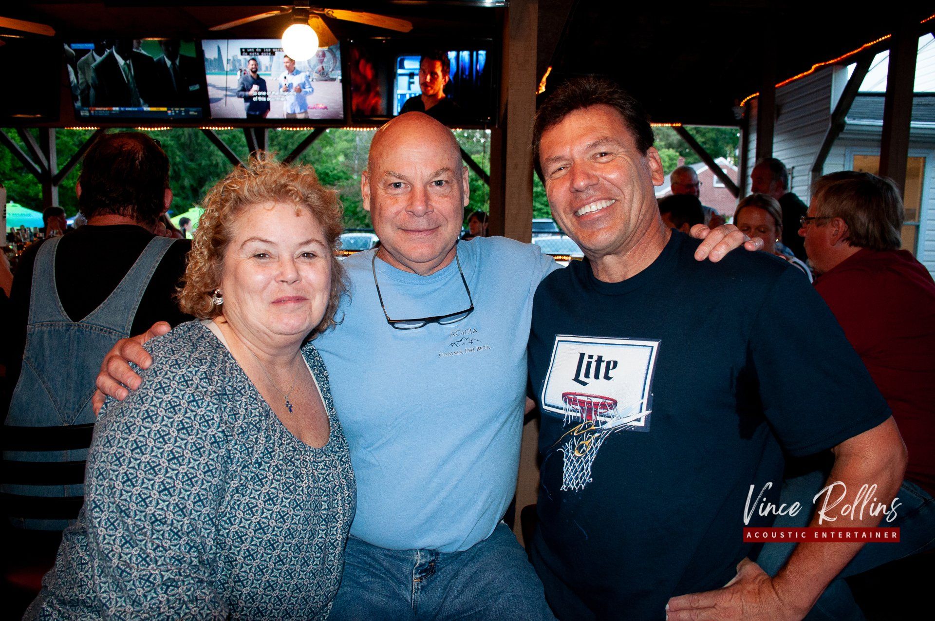 Three smiling people posing together at a lively indoor bar or event.