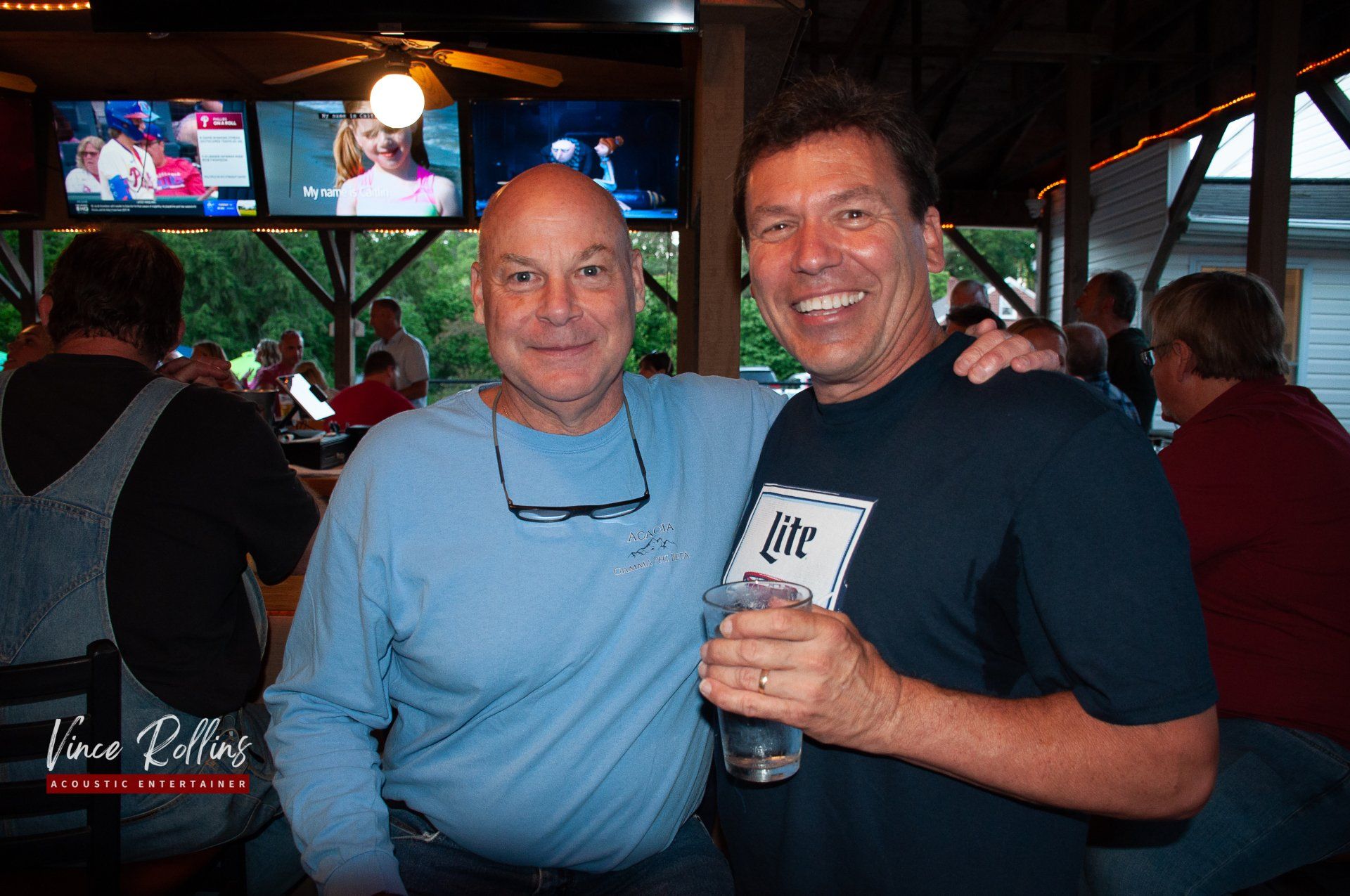 Two men smiling and posing together at a dimly lit bar, one holding a drink.