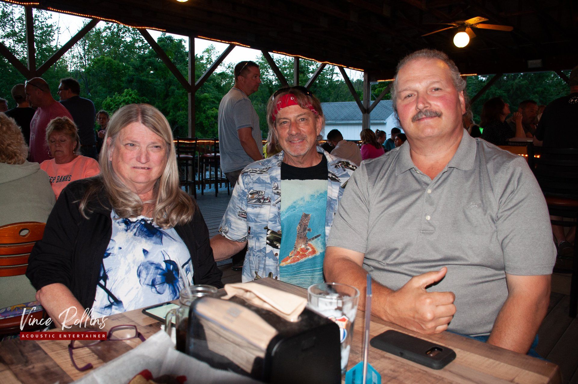 Three people seated at an outdoor table under a covered patio, smiling at the camera.