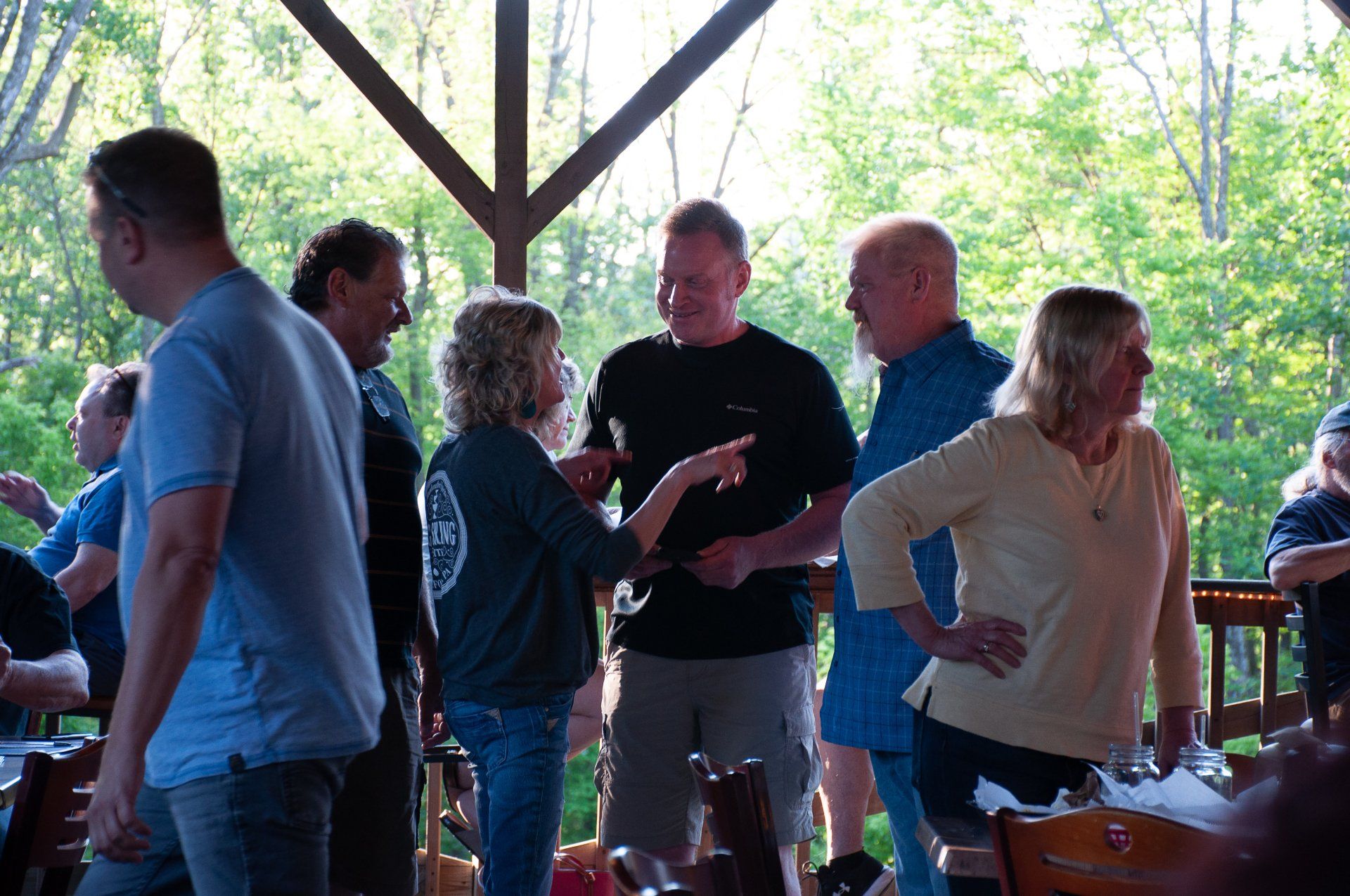 People socializing on a wooden deck in a wooded outdoor pavilion, holding drinks and chatting