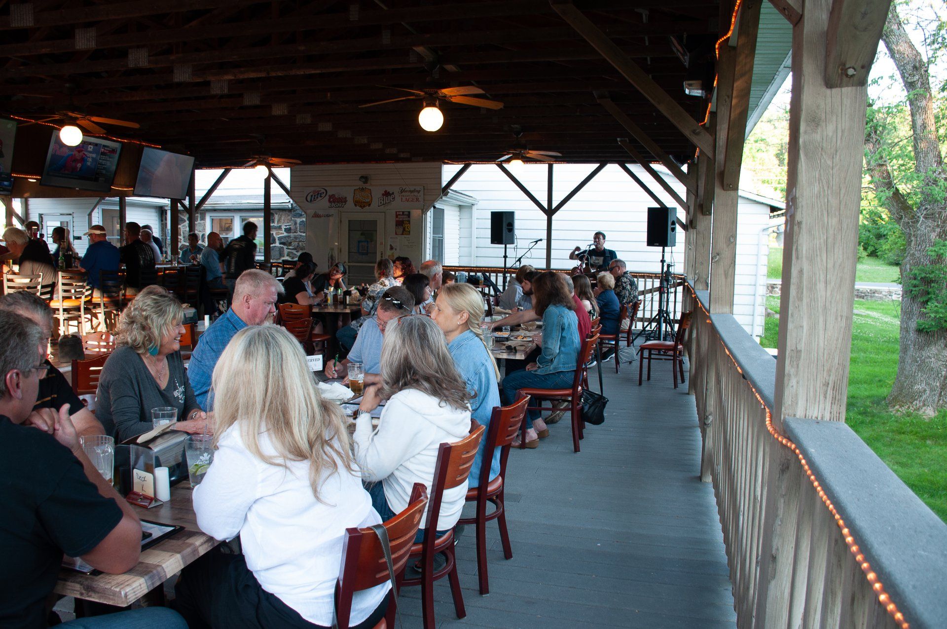 People dining on a covered outdoor patio with wooden tables and chairs, overlooking a grassy area.