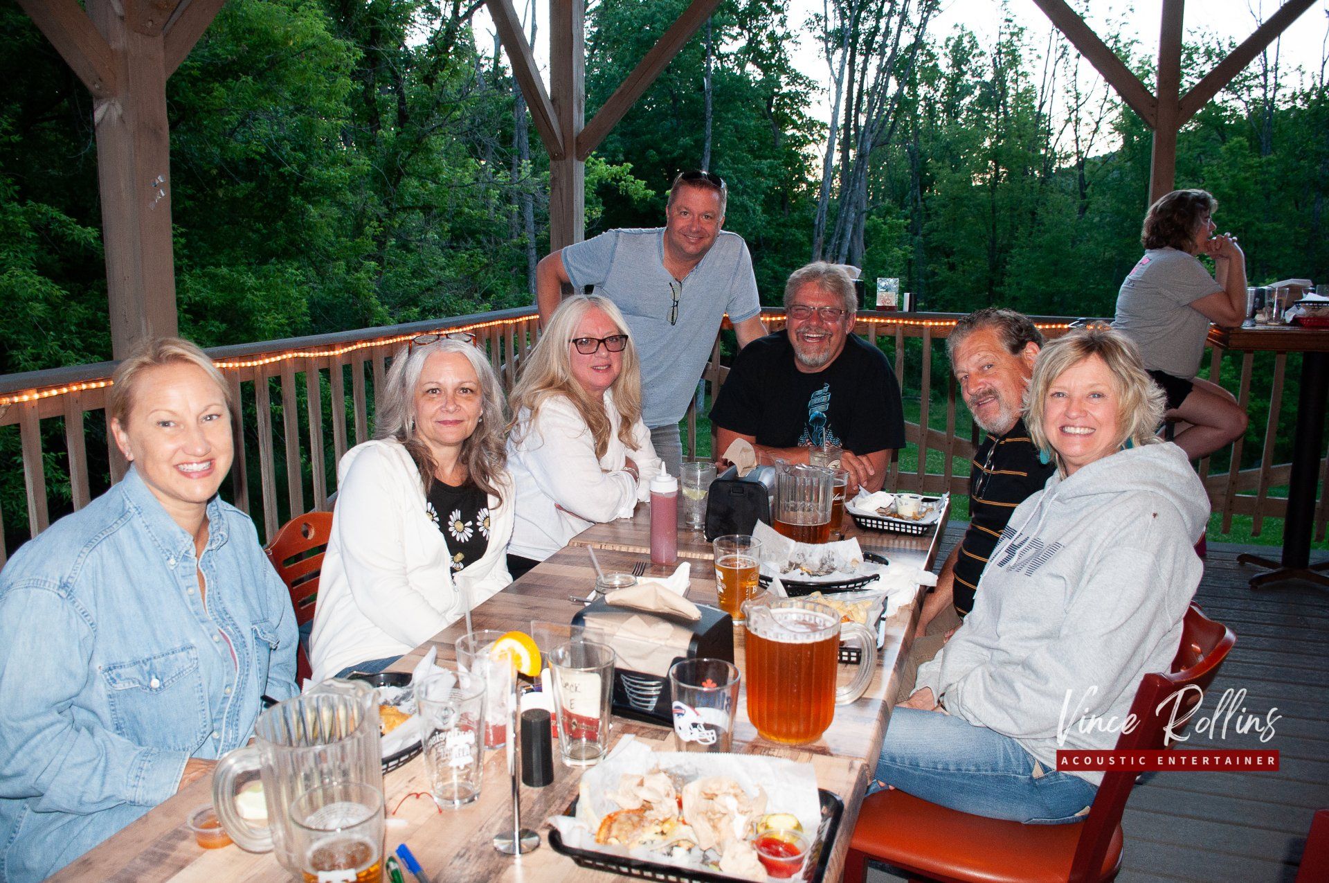 Group of people seated at a table on a wooden deck at dusk, smiling and eating together