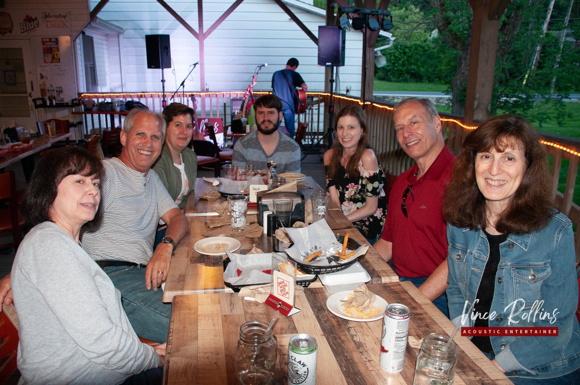 Group of people smiling around a restaurant table, sharing a meal and drinks in a casual indoor setting.