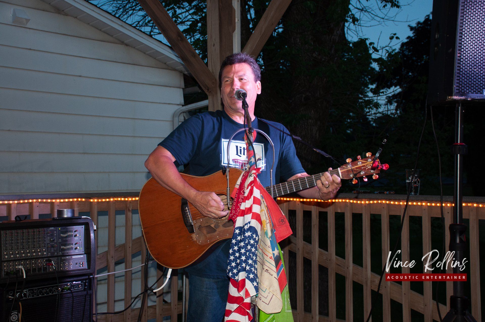 Man playing acoustic guitar and singing on an outdoor deck at dusk, with a microphone and string lights