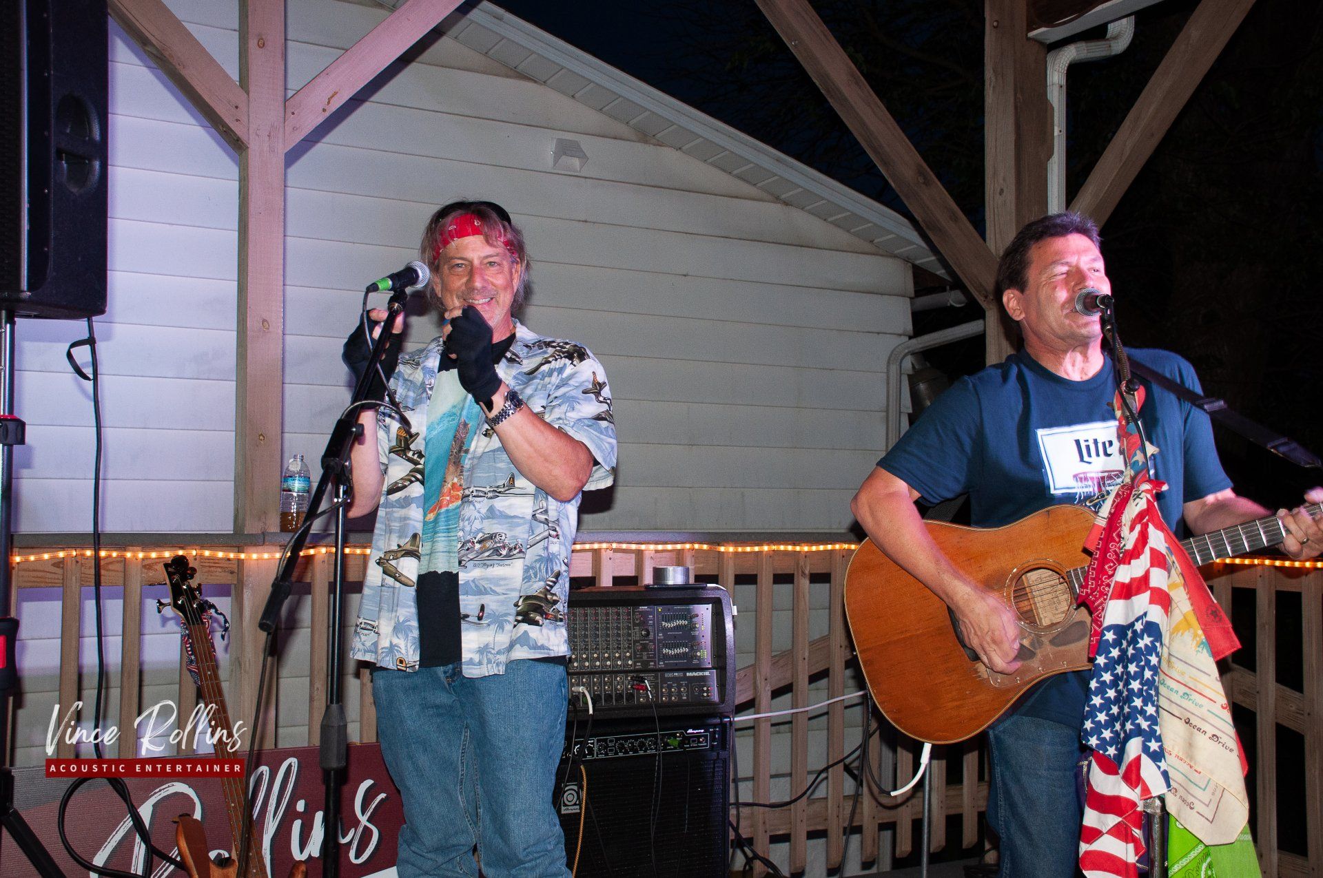 Two musicians performing on a wooden stage, one singing into a mic and one playing guitar.