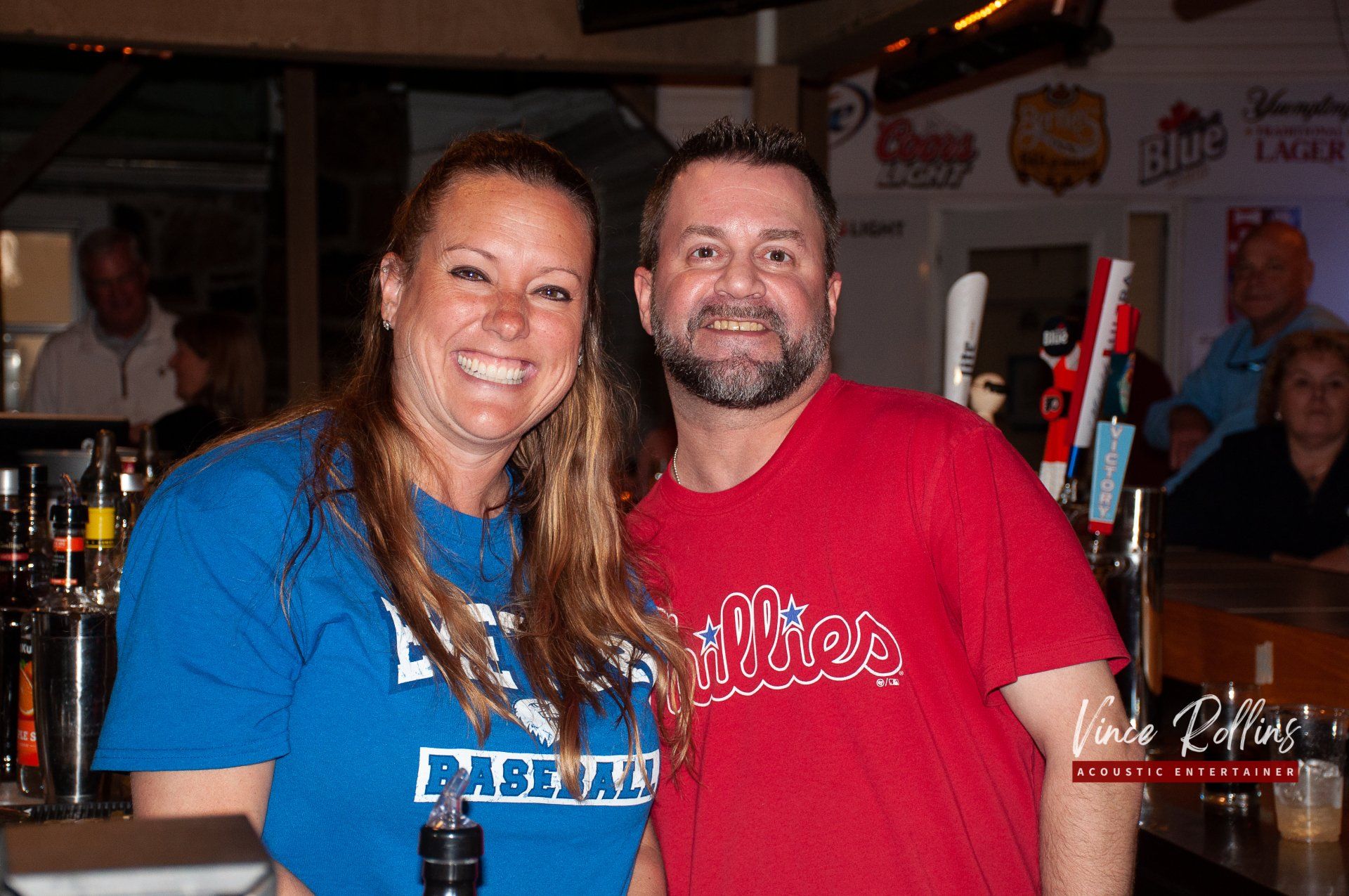 Two smiling people posing together in a dimly lit bar, one in a blue shirt and one in a red shirt.