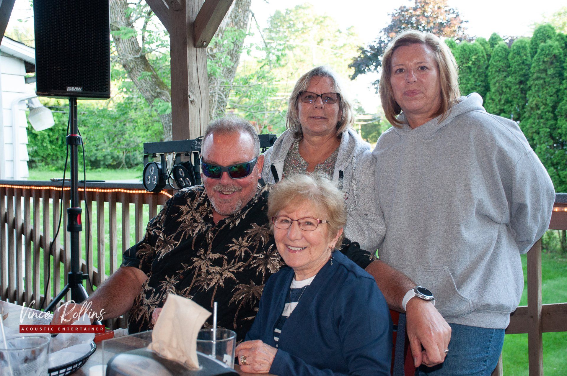 Four people seated and standing on a porch, smiling beside a table with drinks and a speaker.