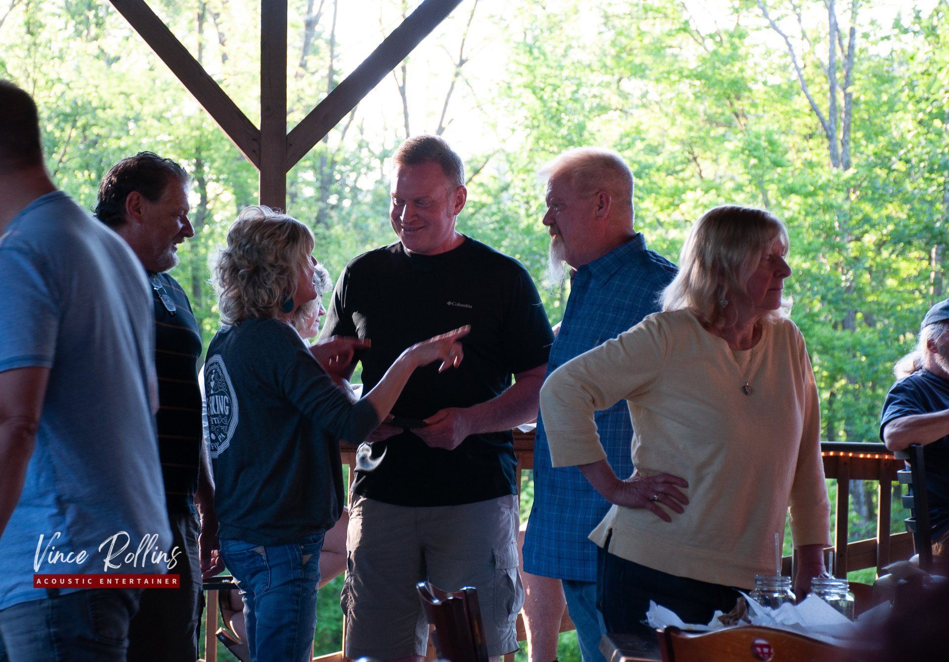 People chatting under a pavilion at an outdoor gathering, with trees and a wooden railing in the background