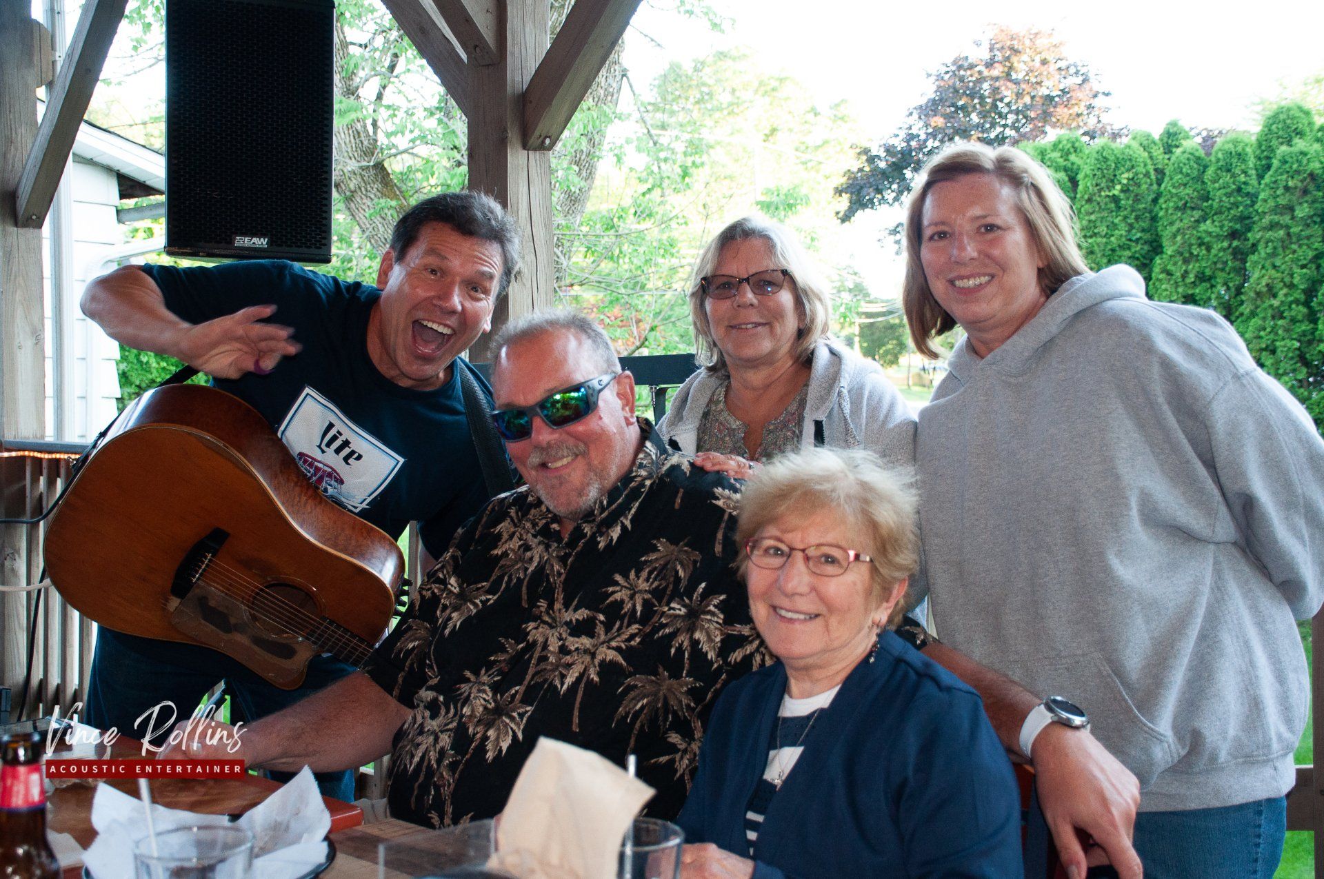 Group of five smiling people gathered outdoors under a pavilion, with one holding a guitar.