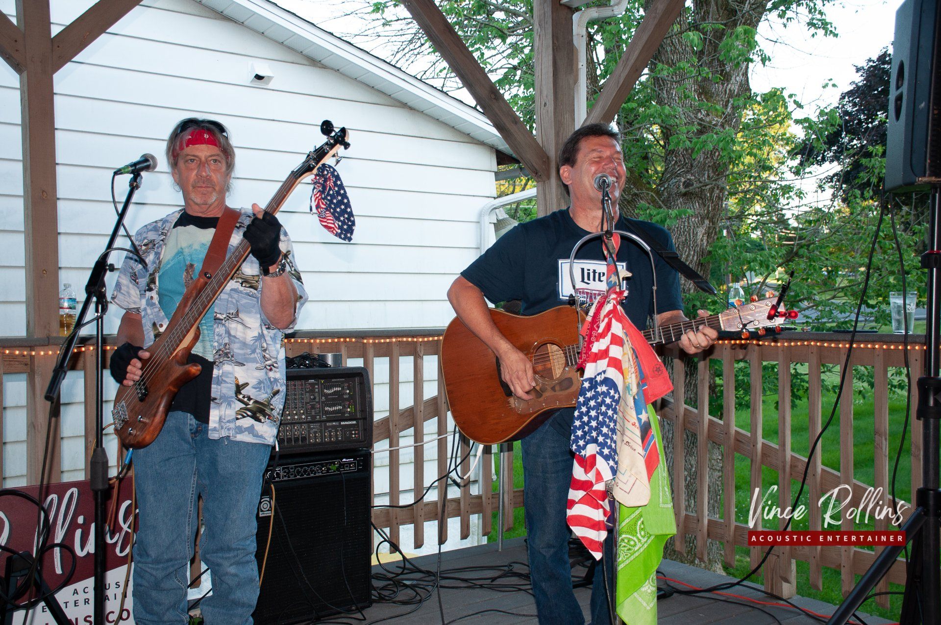 Two musicians performing outdoors, one playing bass and one singing with acoustic guitar and a microphone stand.