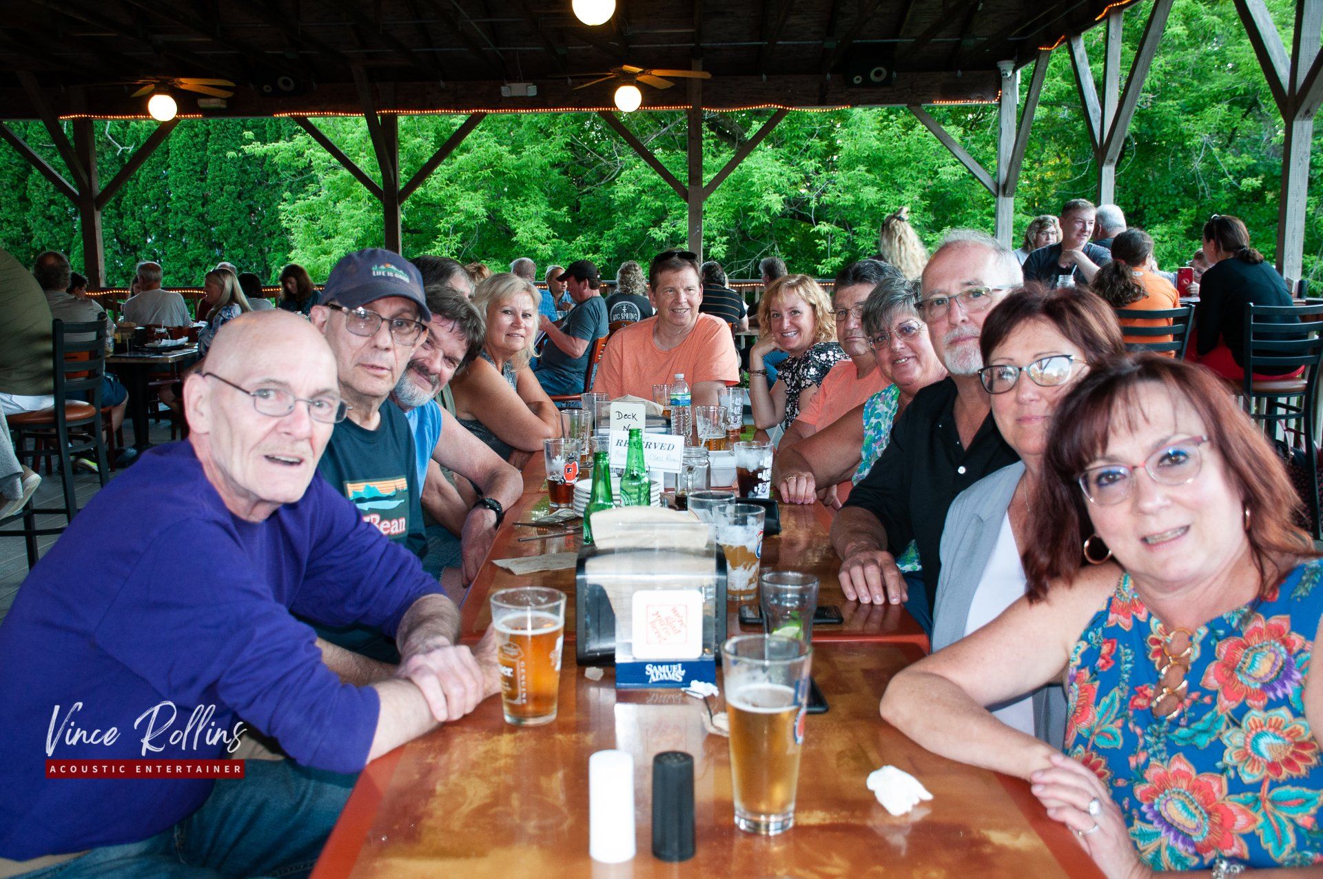 Group seated at a long wooden table in a covered outdoor pavilion, smiling and talking over drinks