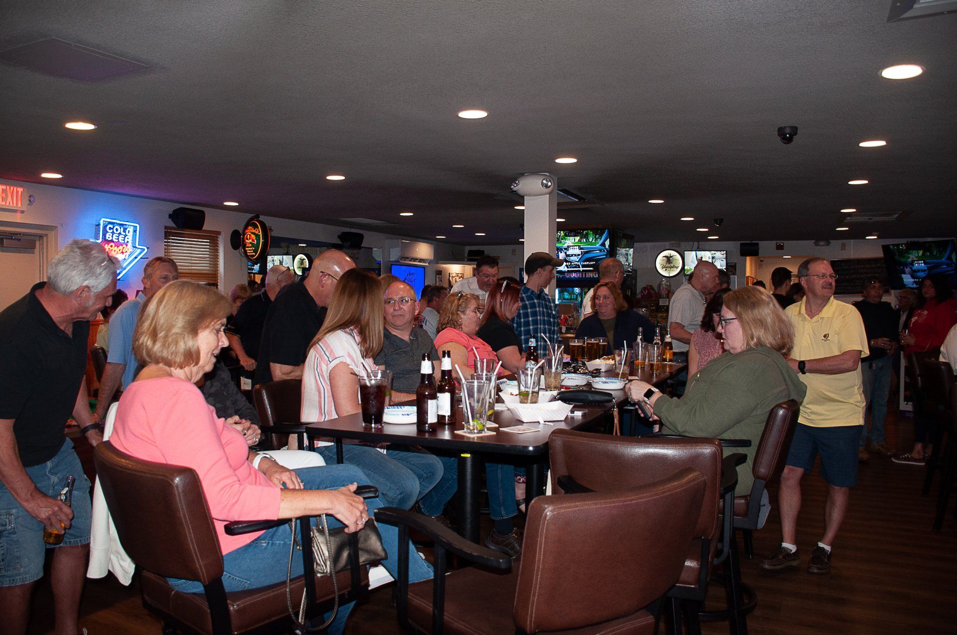 Crowded bar with people seated at tables and standing near the counter, drinks on the tables.