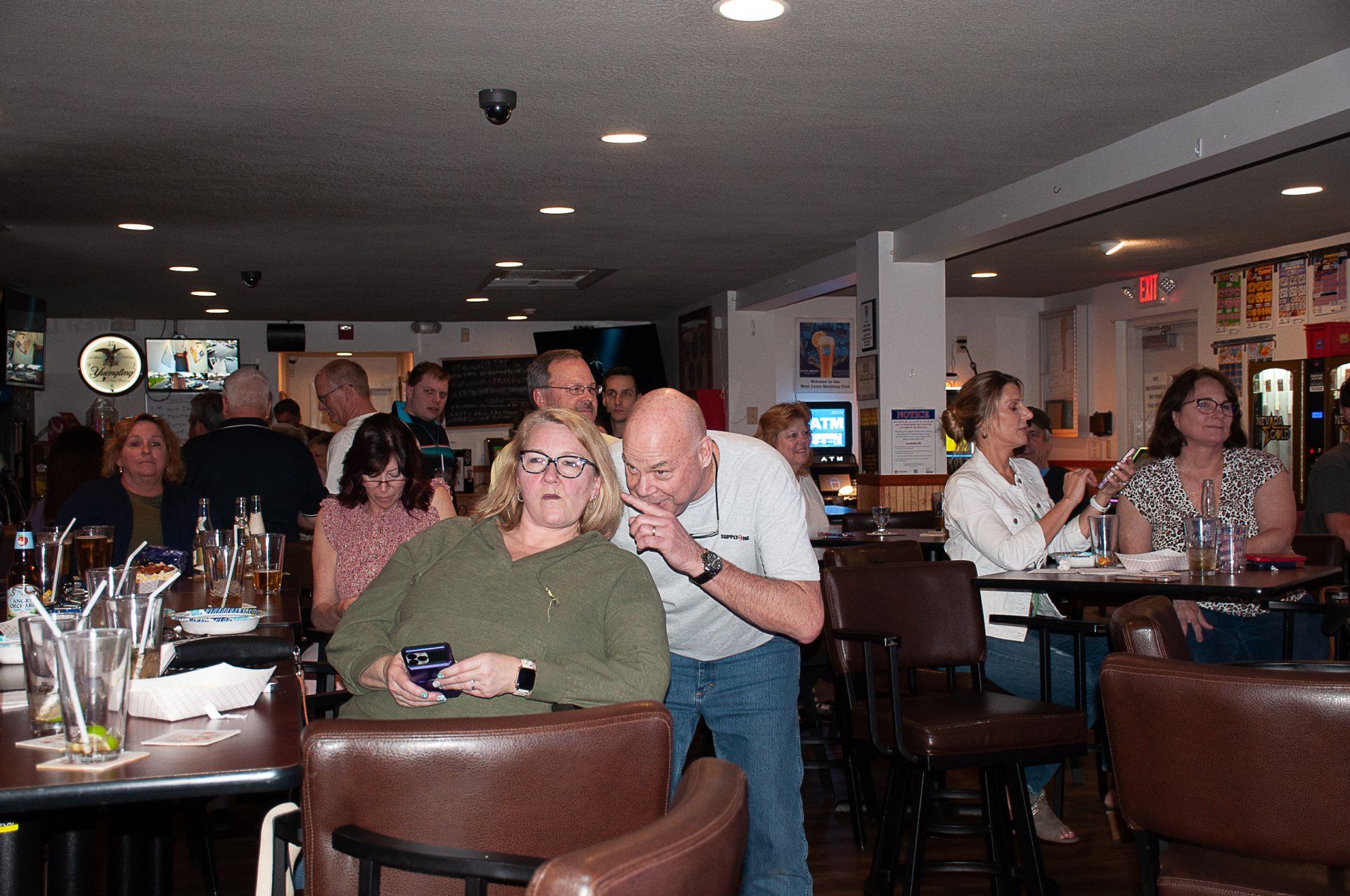 People seated in a dimly lit restaurant or bar, with drinks on tables and one man leaning over a chair.