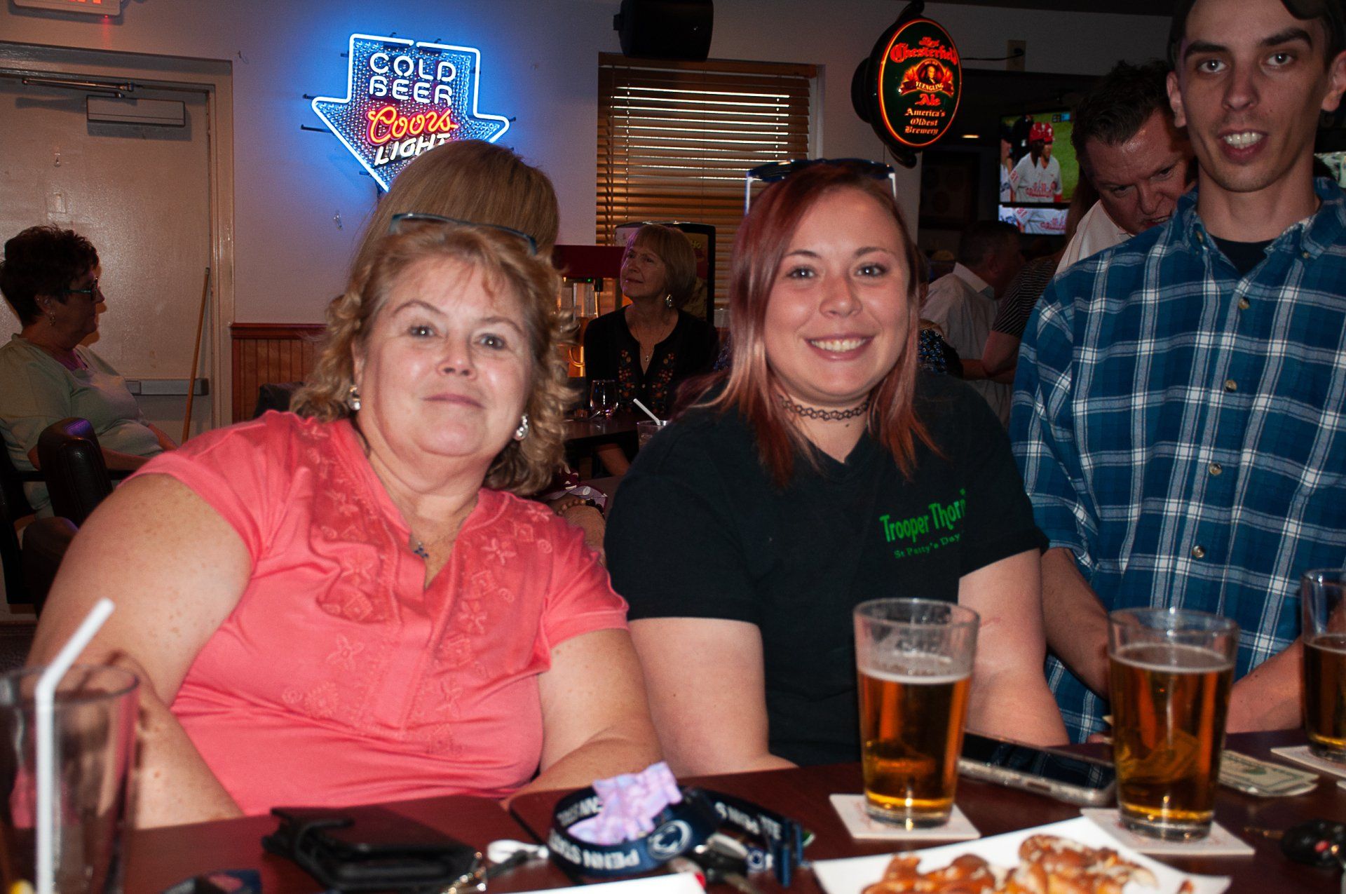 Three people seated at a bar table with drinks and food, smiling in a dimly lit restaurant.