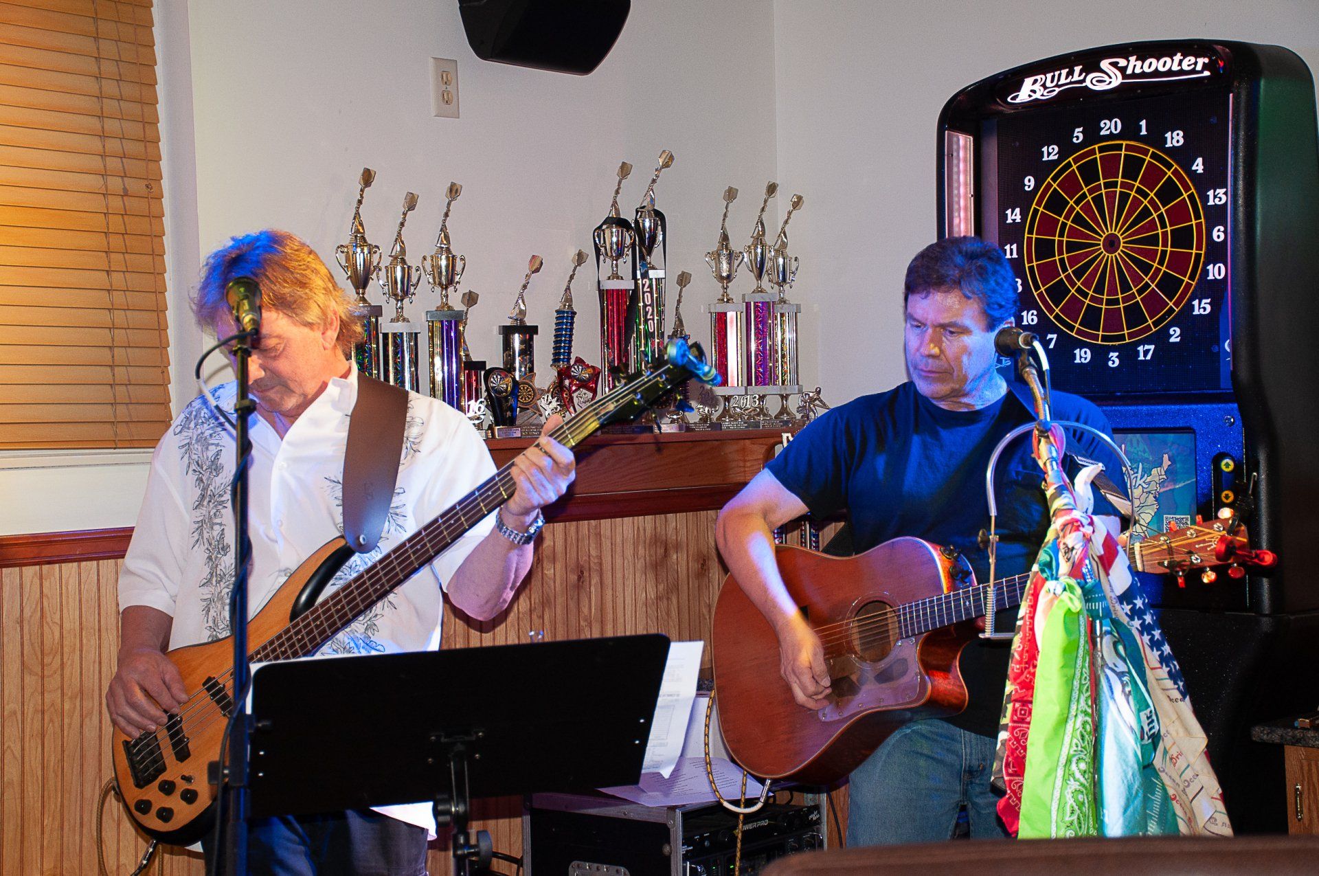 Two musicians playing guitar and singing onstage with a dartboard and trophies in the background